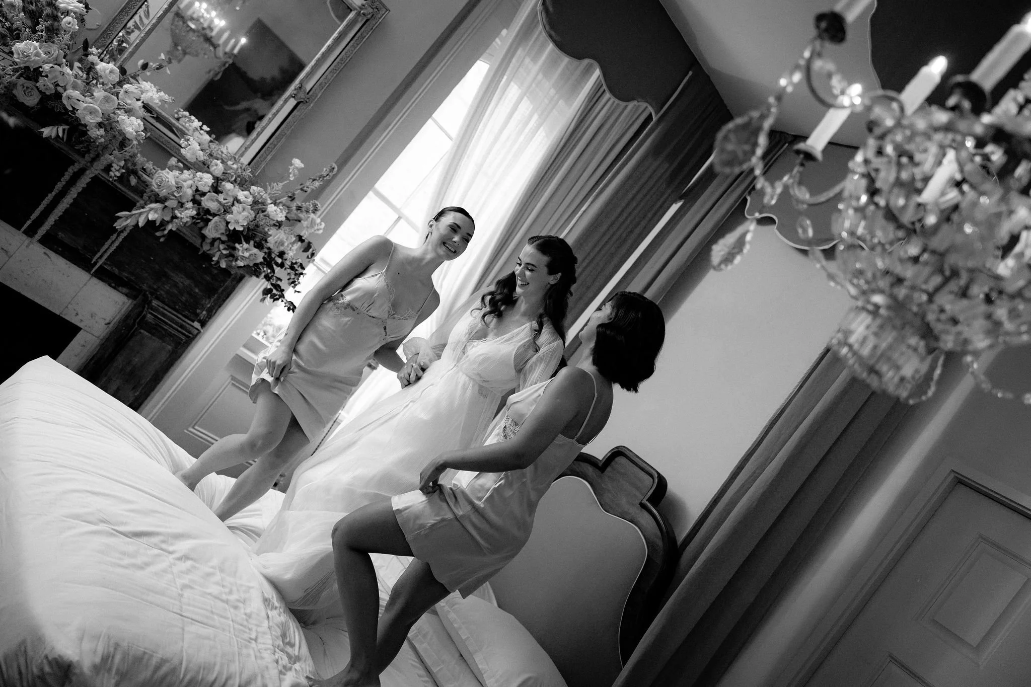 Three women laughing and chatting on a bed in a well-decorated bedroom with a chandelier, large window, and floral arrangements.