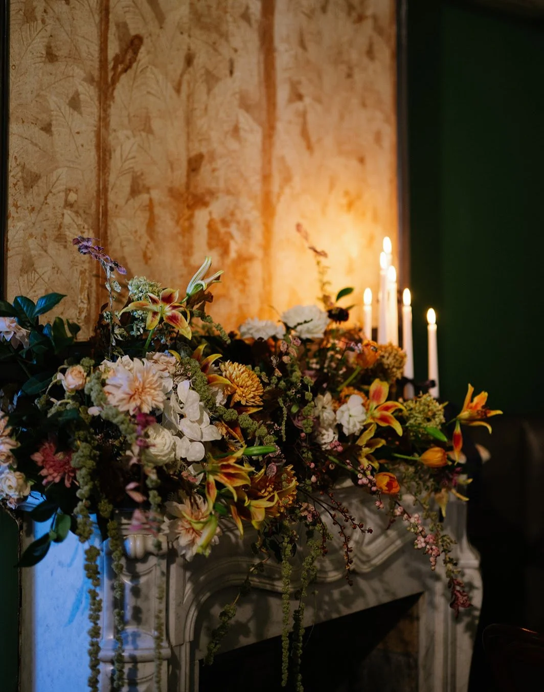 Floral arrangement with pink, orange, and white flowers on a fireplace mantel, lit by candlelight, against a textured warm wall with dark green on the side.