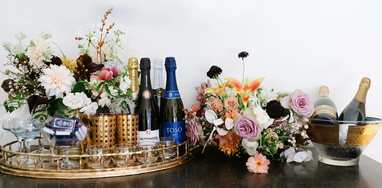 A tray containing multiple bottles of sparkling wine, glasses, and candles, surrounded by a floral arrangement of pink, white, and purple flowers, with a champagne bucket on the right.