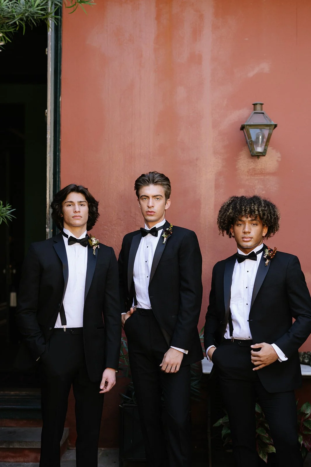 Three young men wearing black tuxedos and bow ties pose in front of a reddish wall, each with a boutonniere on their lapel, standing confidently with serious expressions.