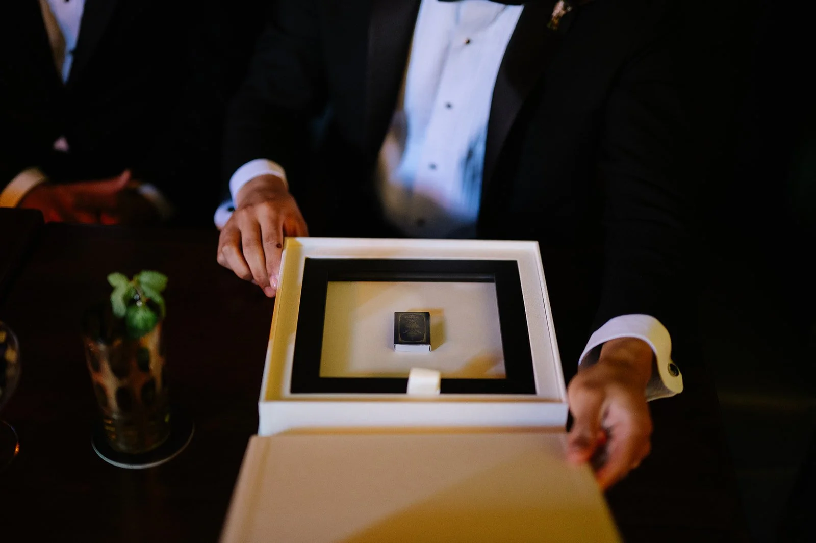 A man in a tuxedo opening a white box revealing a black box inside, on a dark wooden table with a glass of drink and a plant on the side.