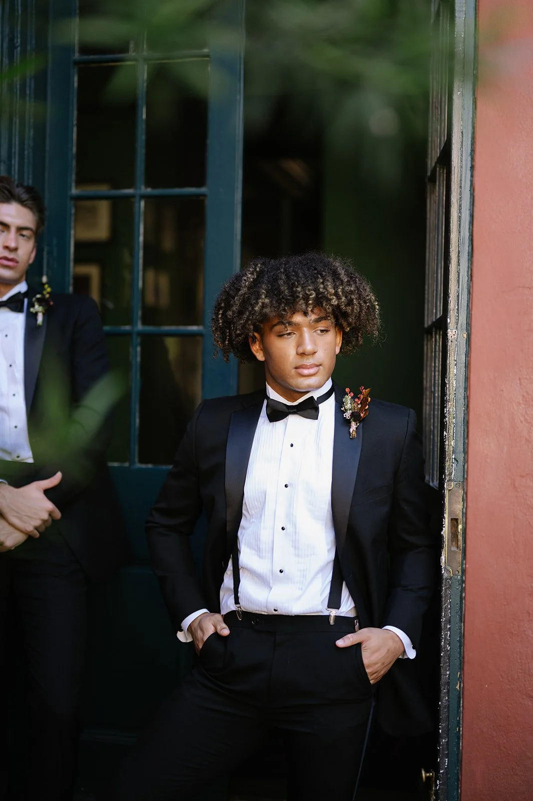 Two men in black tuxedos with white shirts and black bow ties standing near a door, one leaning against the door frame with hands in pockets and the other standing in the background with arms crossed.
