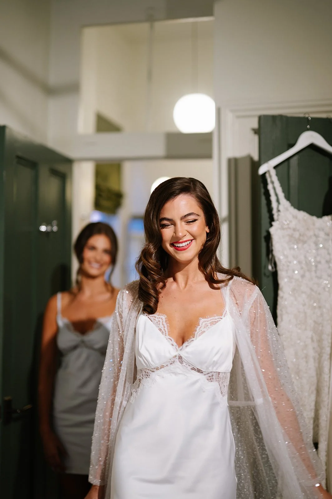 A woman in a white wedding dress smiling with her eyes closed, another woman in a grey slip dress standing behind her, and a wedding dress hanging on a hook in a fitting room.