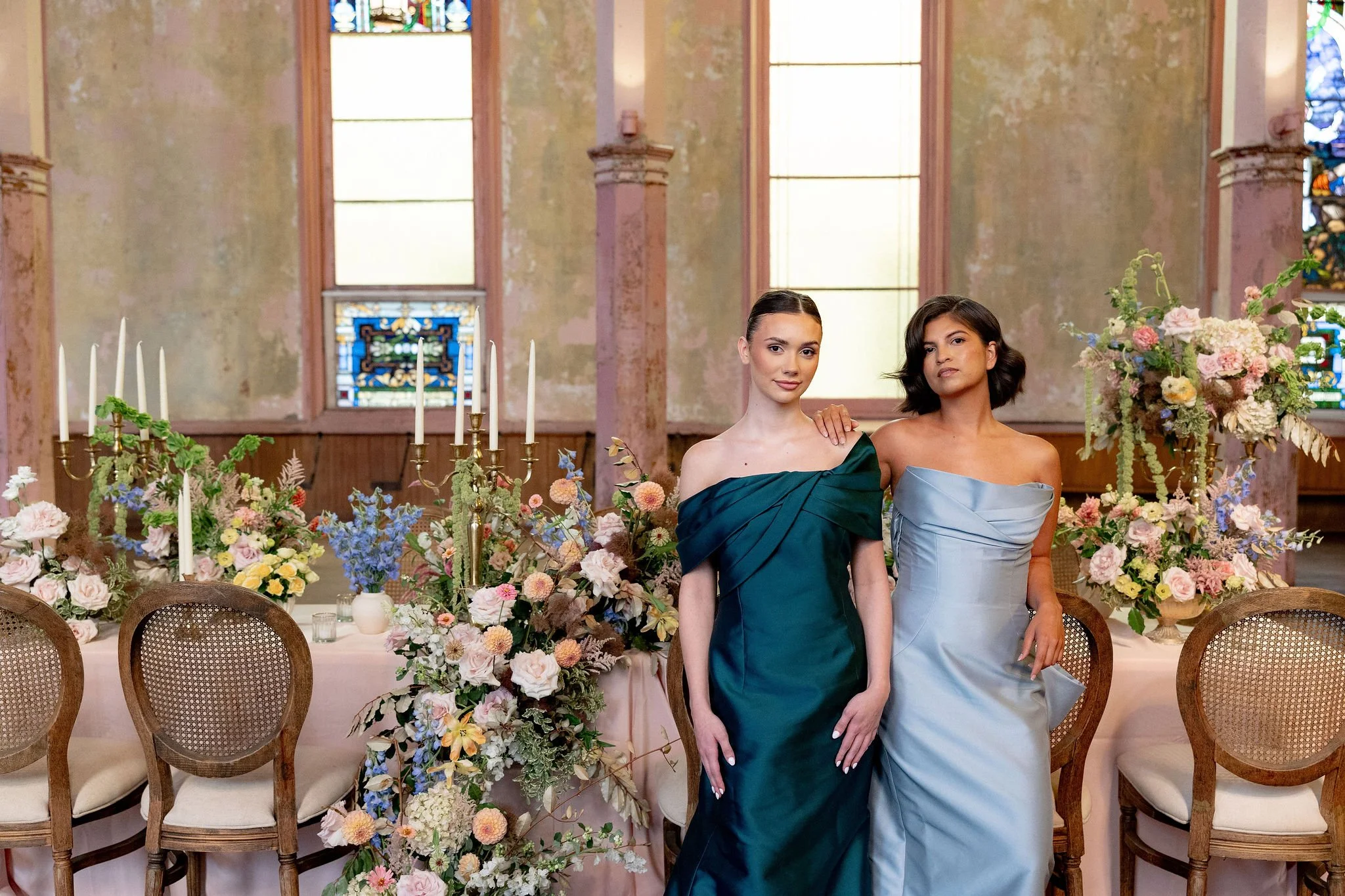 Two women in formal dresses standing in front of a decorated table with flowers and candles inside a church or chapel.