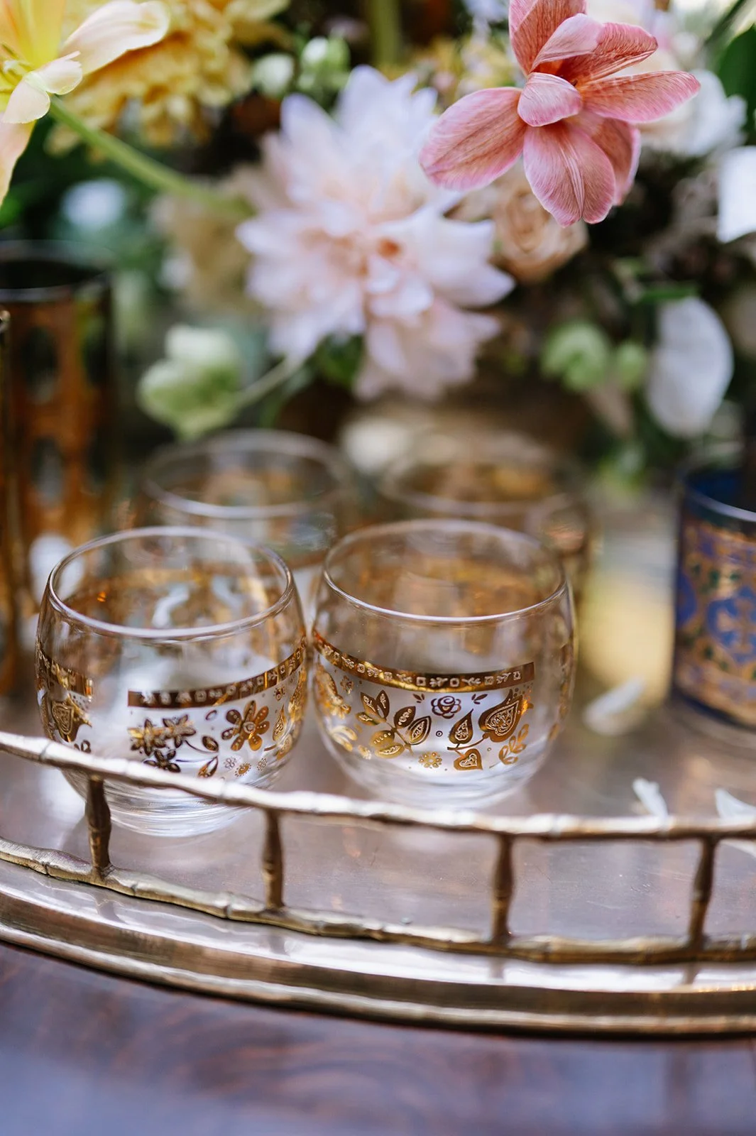 Elegant glass cups with gold floral patterns on a silver tray, surrounded by pink and white flowers.