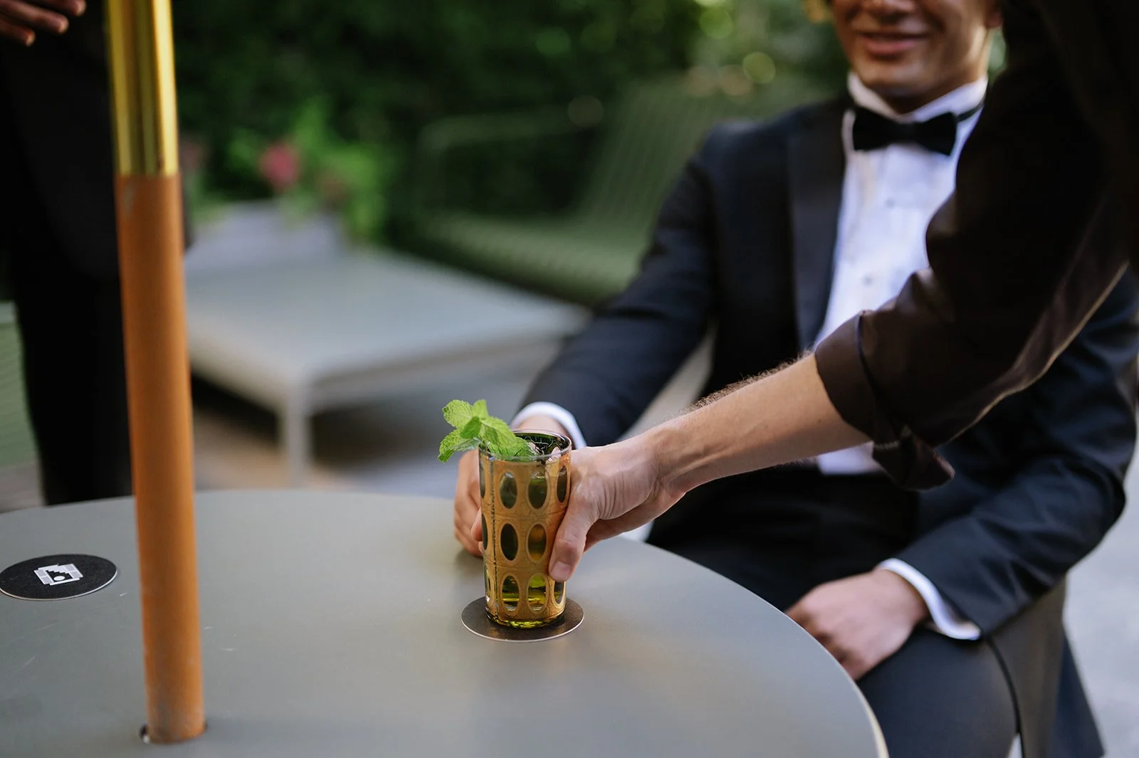 A waiter serving a drink with mint leaves to a man in a tuxedo sitting at an outdoor table.
