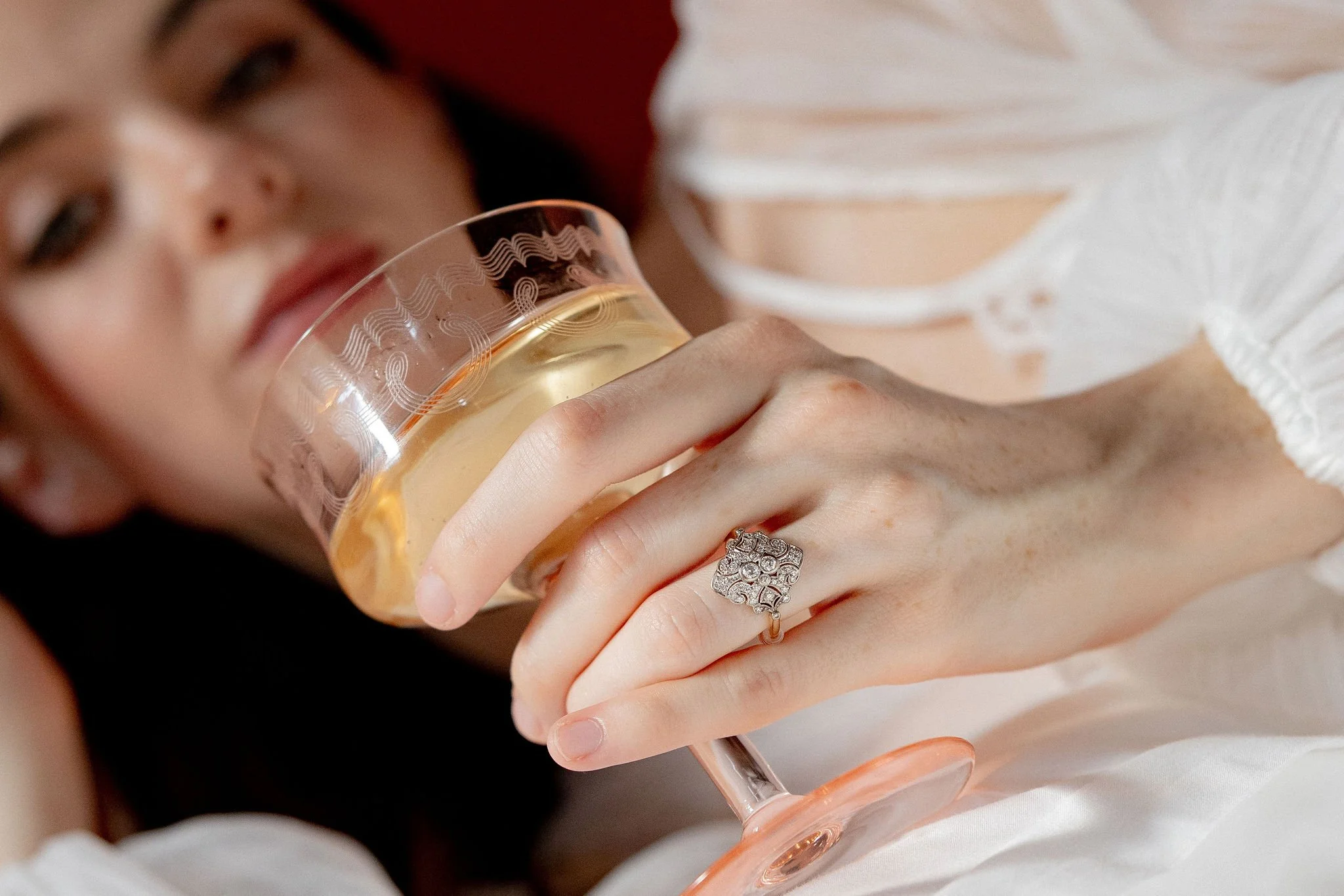 A woman lying down, holding a glass of white wine or champagne, wearing a silver ring with intricate design, dressed in white, with her face slightly blurred in the background.