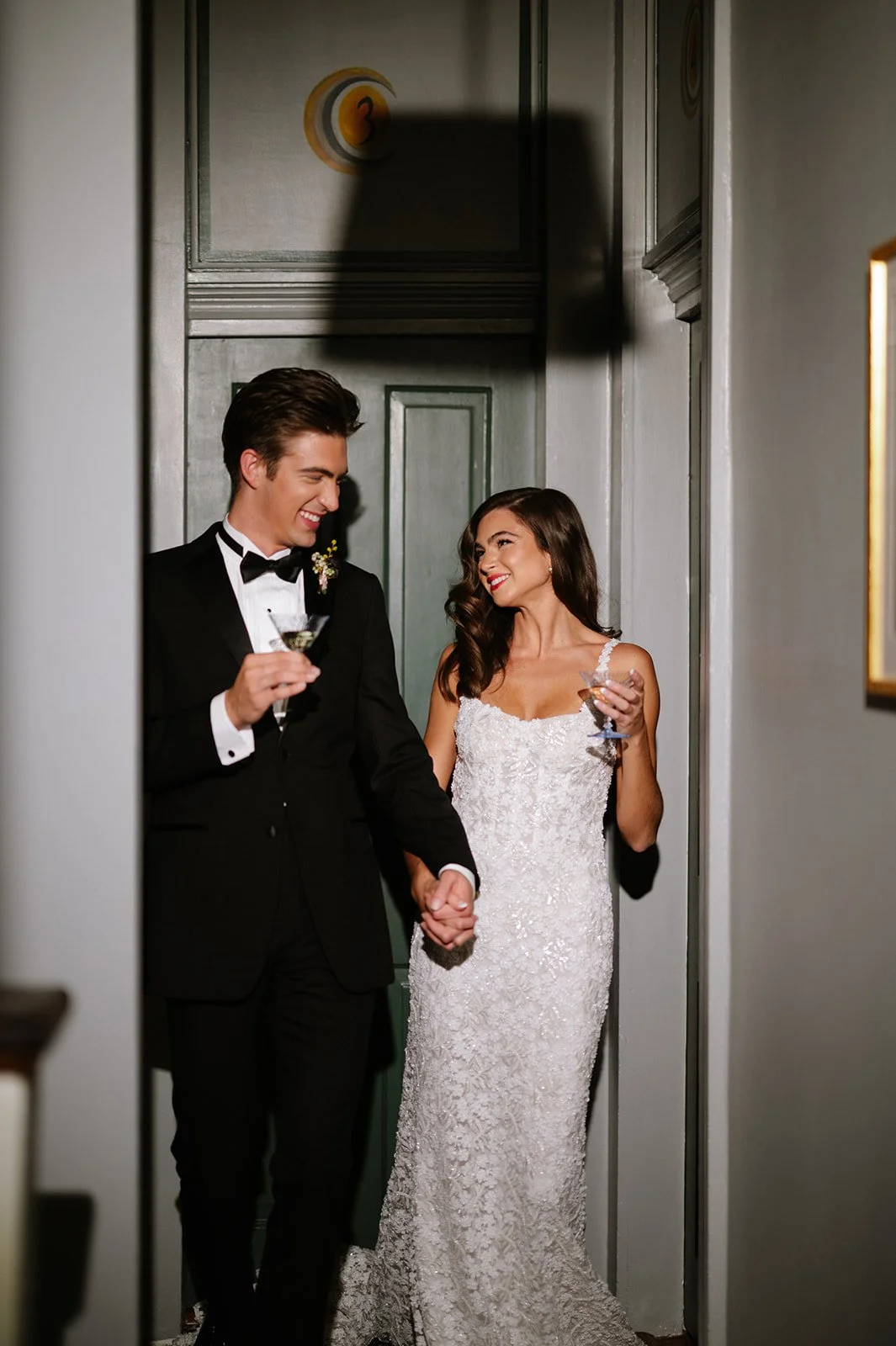 A newlywed couple, dressed in wedding attire, holding hands and enjoying drinks while peeking out from a hallway.
