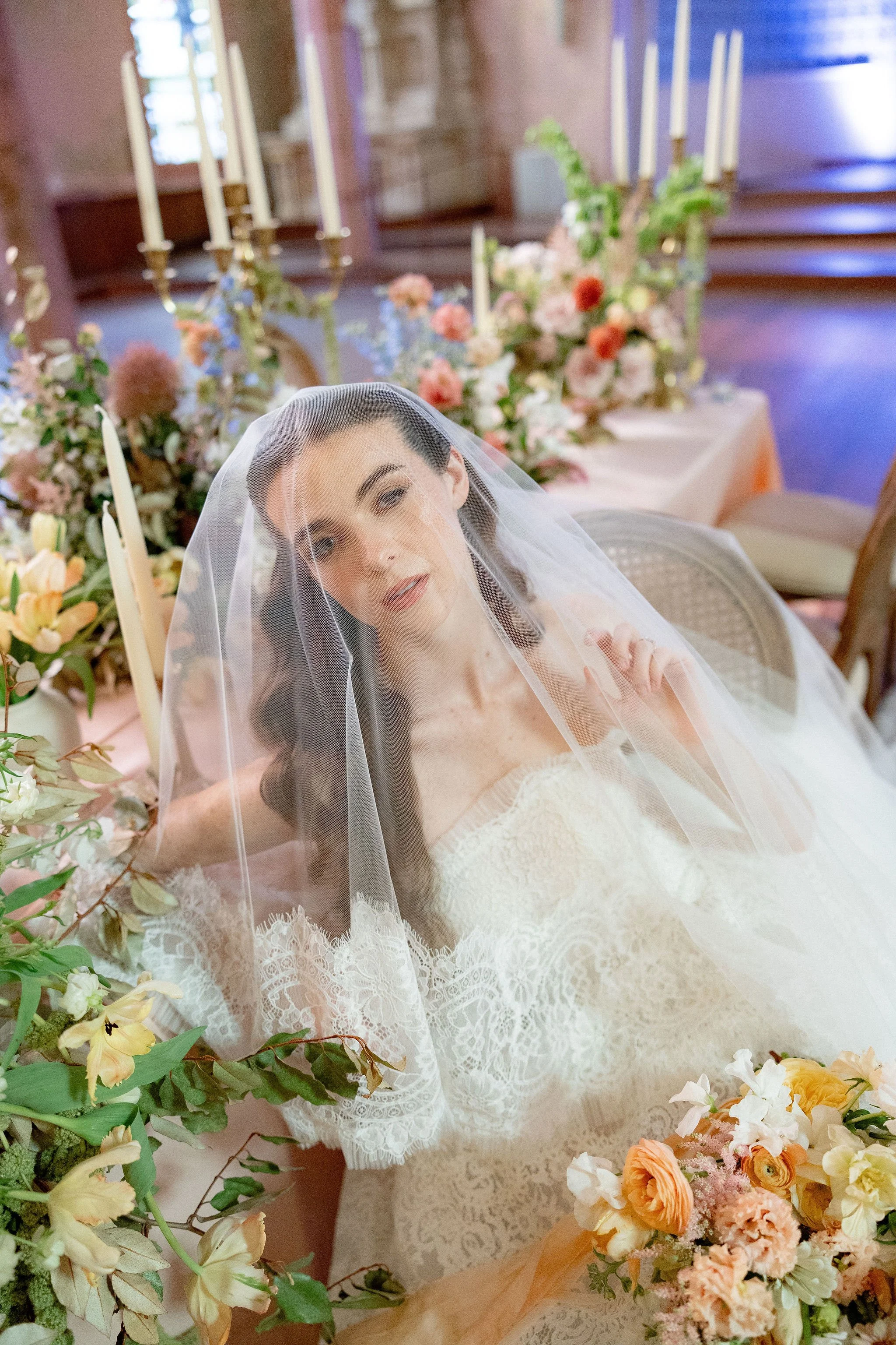 A bride in a wedding dress and veil sitting at a decorated table with flowers, candles, and candelabras.
