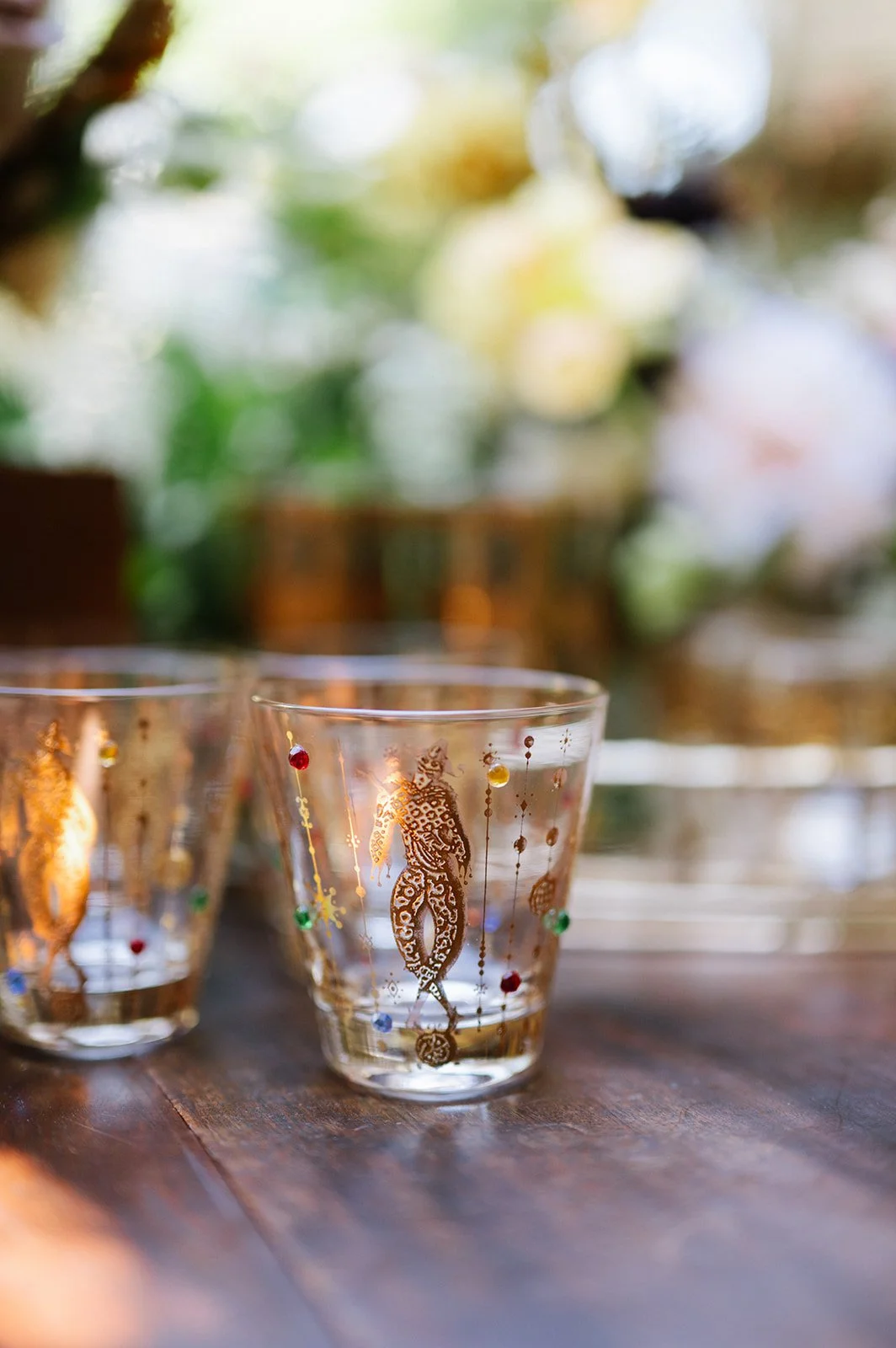Close-up of clear glass candle holder decorated with a gold mermaid design and colorful rhinestones, placed on a wooden surface with blurred background of greenery and holiday decorations.