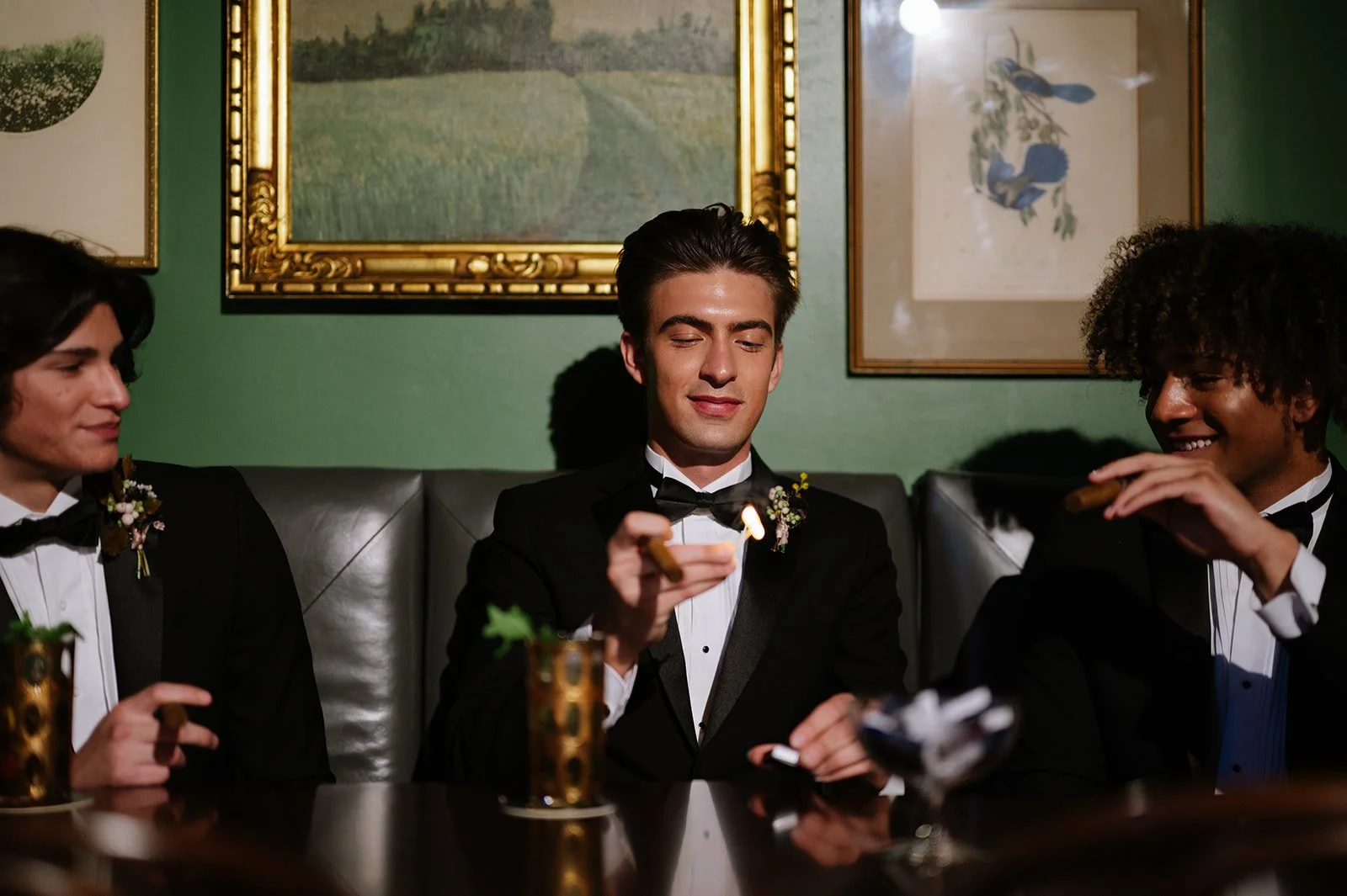 Three men in tuxedos sitting on a green wall background, with framed paintings, at a formal gathering. The man in the middle is holding a lit cigar, while the man on the right is also holding a cigar and smiling. The man on the left looks on. Two drinks in gold holders are on the table.