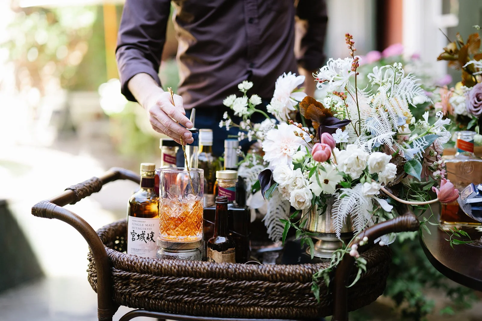 A person pouring a drink into a glass on a wicker table surrounded by bottles and a large floral arrangement