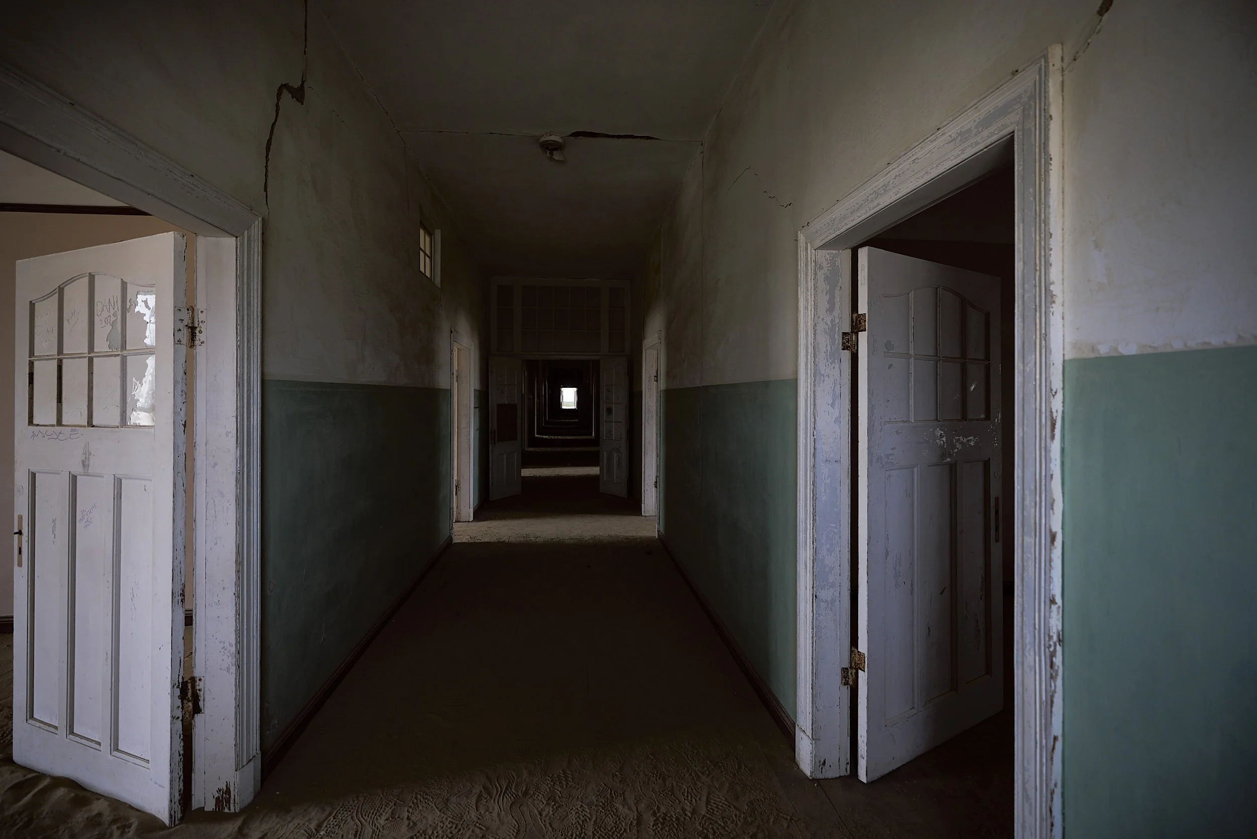 An interior shot of an abandoned building with multiple open doors, peeling paint, cracked walls, and a long corridor leading to a light-filled space in the distance.
