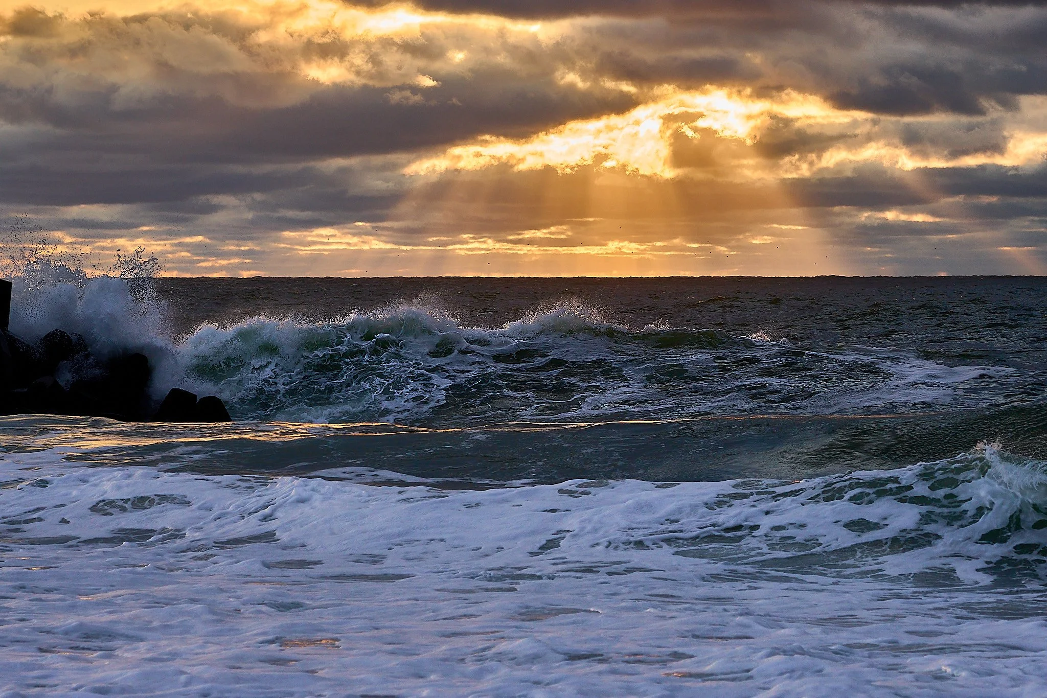 Ocean waves crashing against rocks with a cloudy sky and rays of sunlight breaking through at sunset.