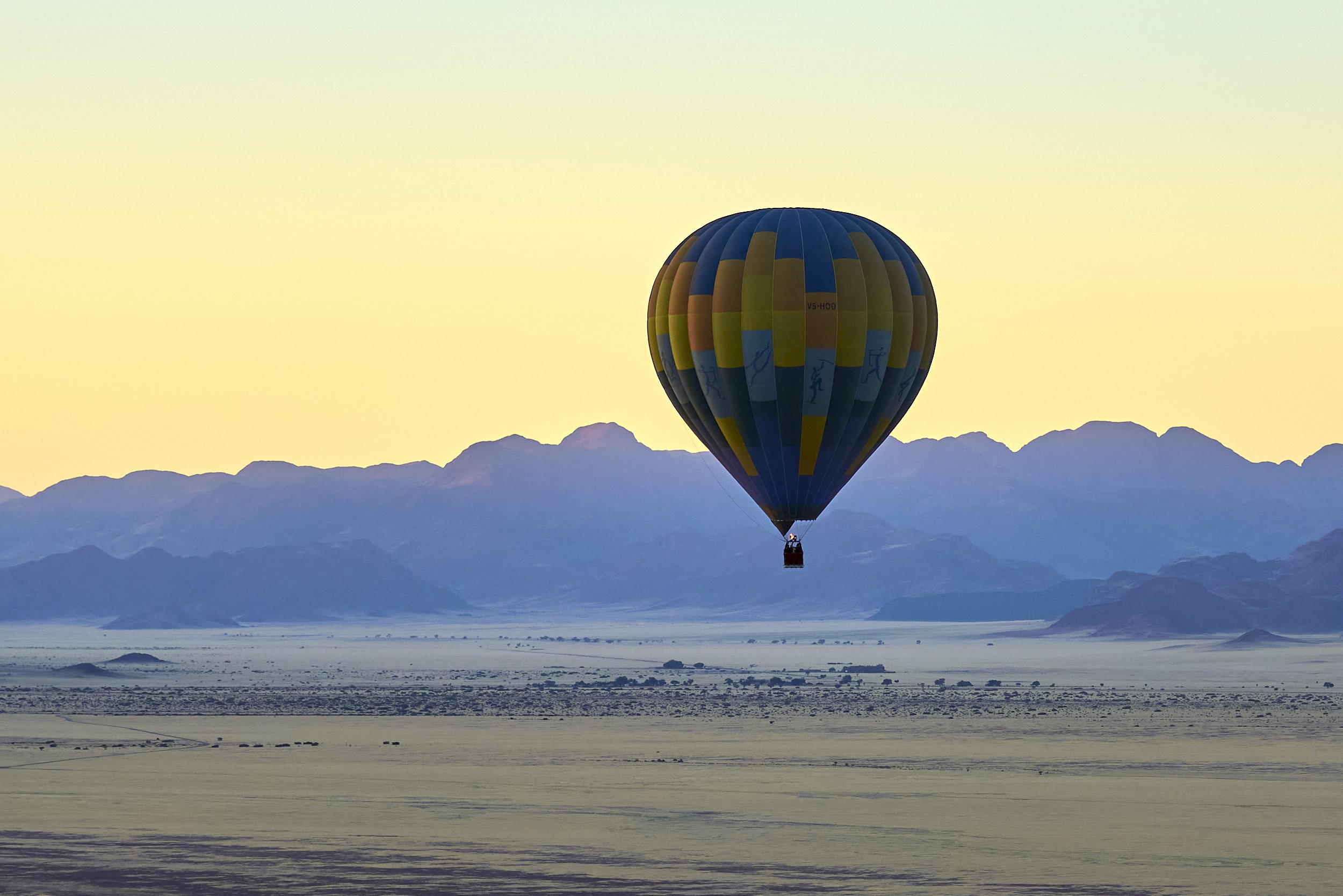 Hot air balloon flying over a flat landscape with mountains in the background during sunset or sunrise.