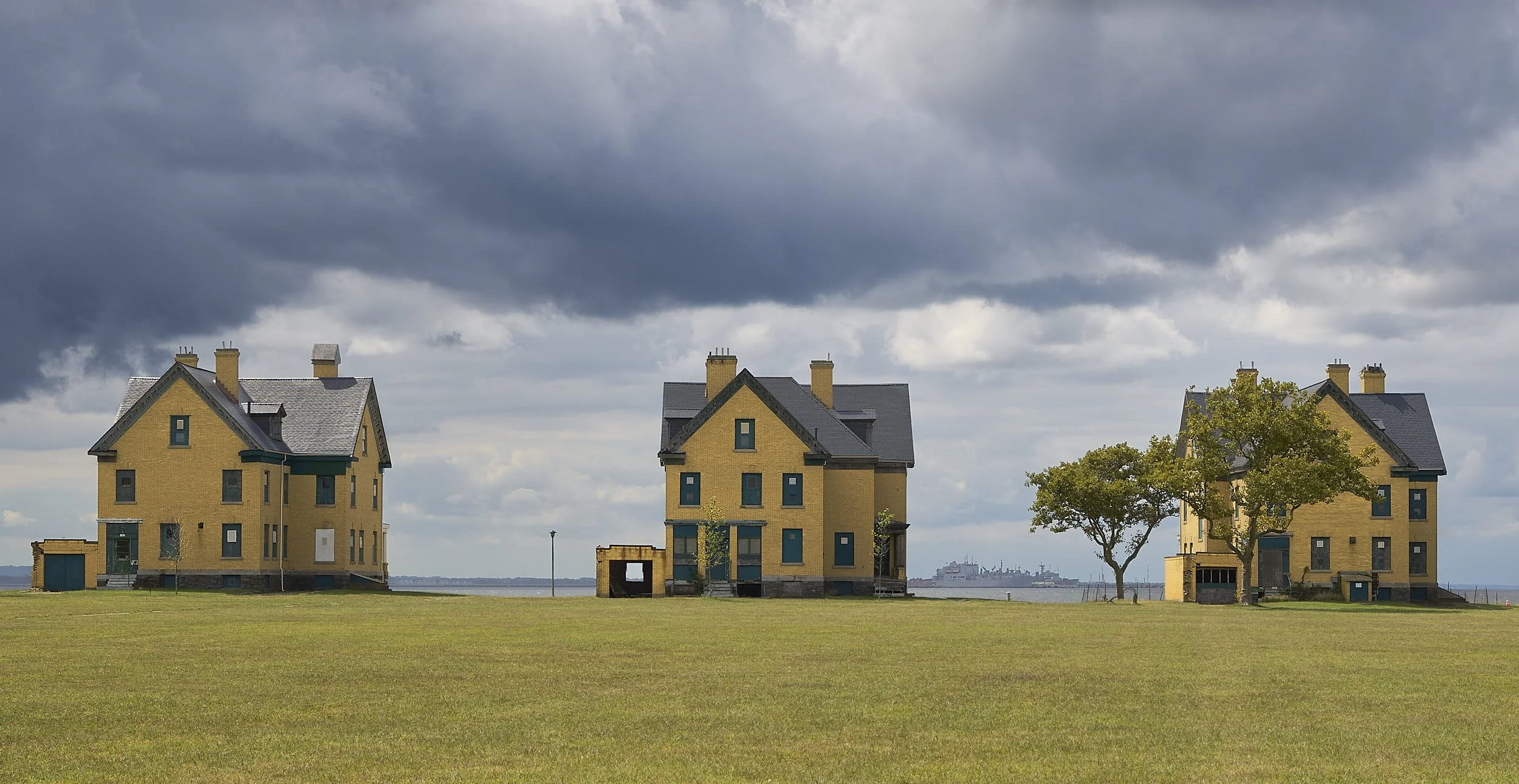 Three yellow wooden houses with dark roofs, set on a grassy field under a cloudy sky, with a body of water and ships in the distance.