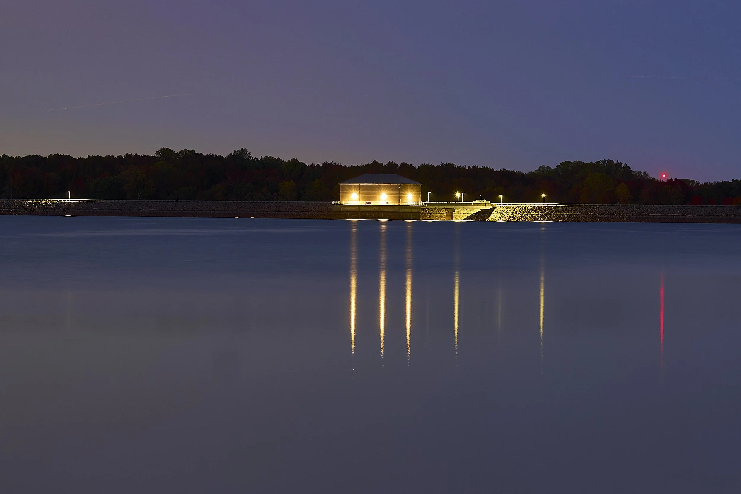 Nighttime view of a water reservoir with a dam, with bright lights illuminating the structure and reflecting on the water surface.