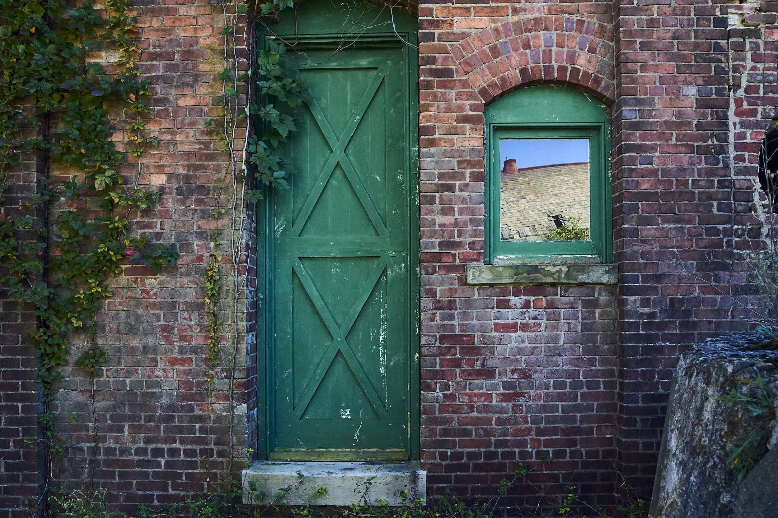 Brick building with a green wooden door and a small green window, ivy growing on the wall, and a reflection of a roof in the window.