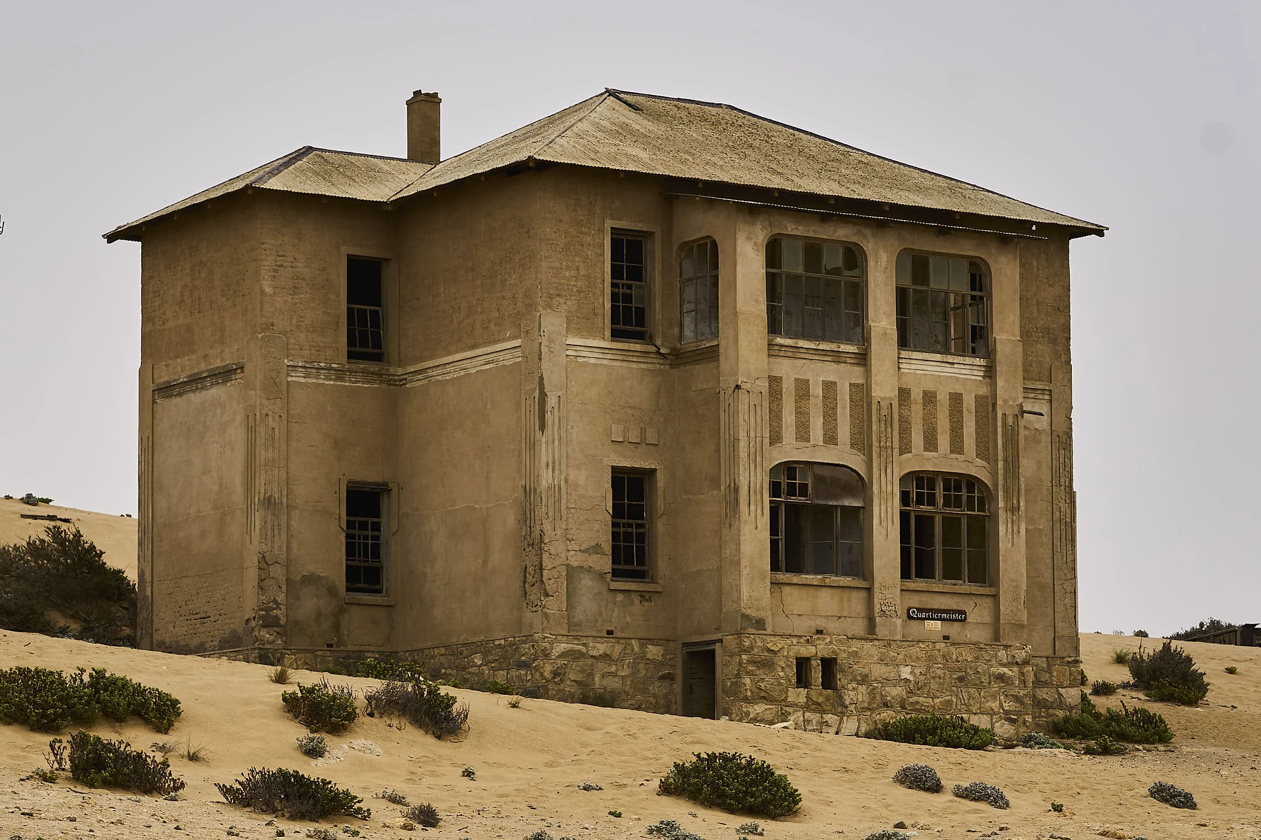An old abandoned house with a beige stucco exterior, multiple windows, and a stone foundation, situated on sandy terrain with sparse bushes.