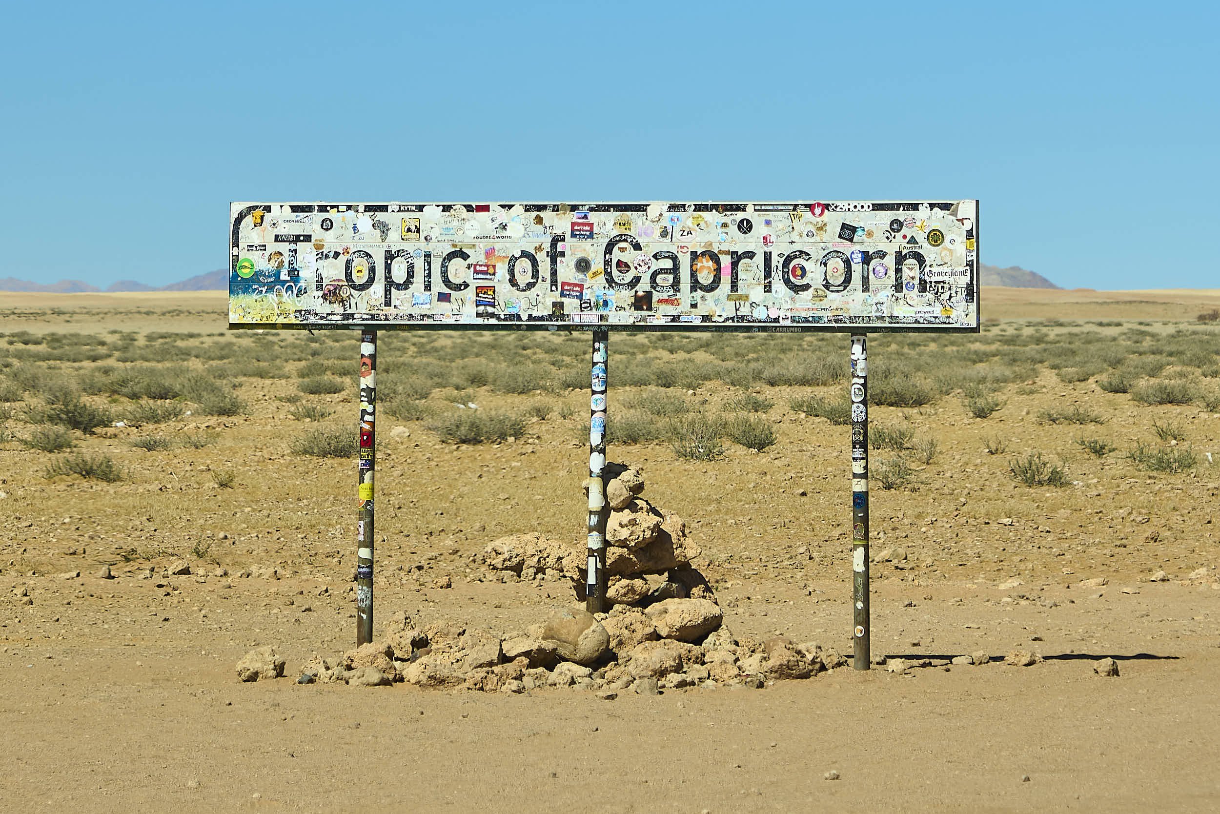 Sign that reads 'Tipic of Cappicorn', covered with many stickers, in a desert landscape with sparse vegetation and mountains in the distance under a clear blue sky.