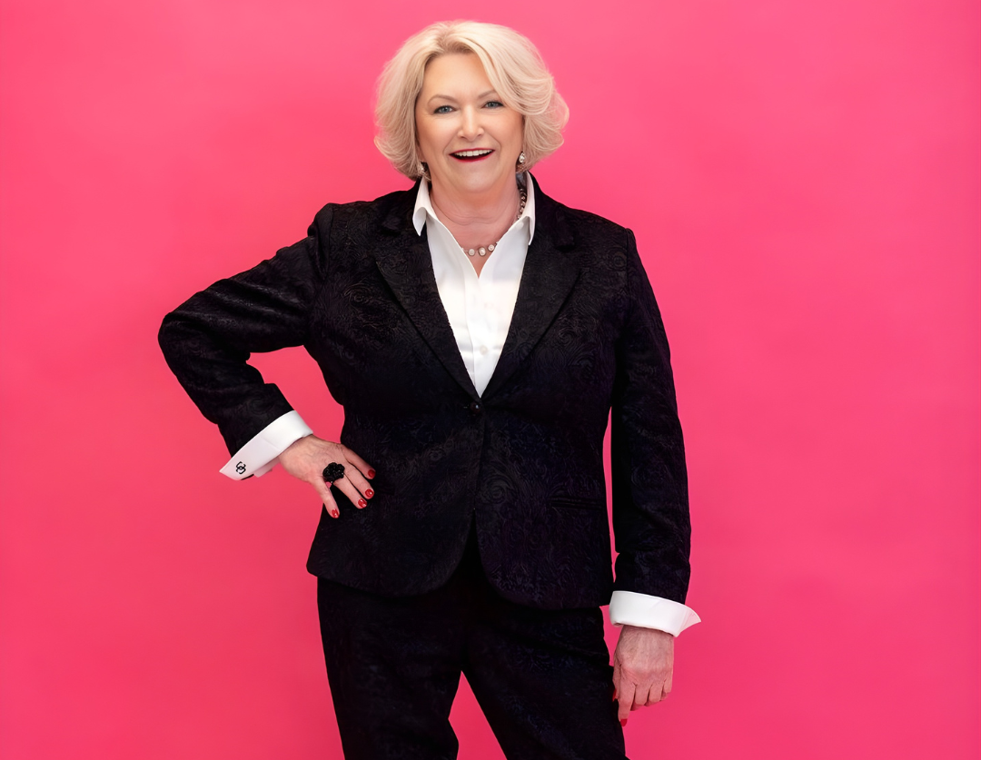 A smiling woman with short blonde hair, wearing a black suit jacket with intricate patterns, a white dress shirt, and black pants, standing against a pink background.