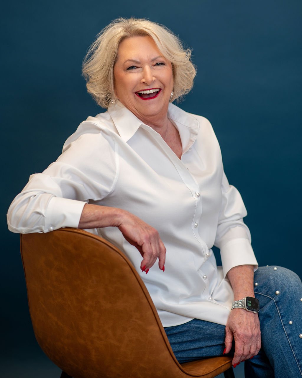 An older woman with blonde, wavy hair, smiling, wearing a white button-up shirt, sitting on a brown chair against a dark blue background.