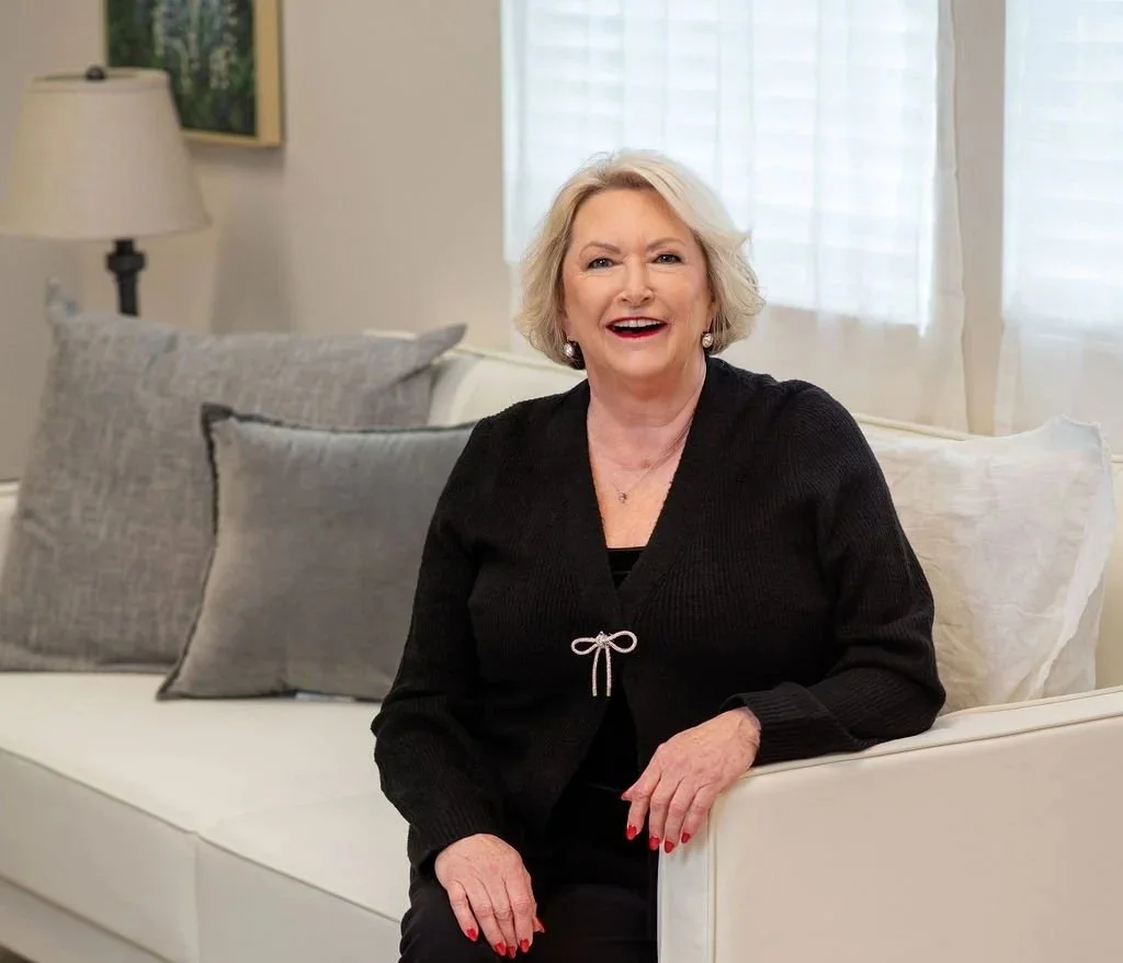 An older woman with blonde hair, smiling, sitting on a white couch with gray pillows in a bright, well-lit living room.