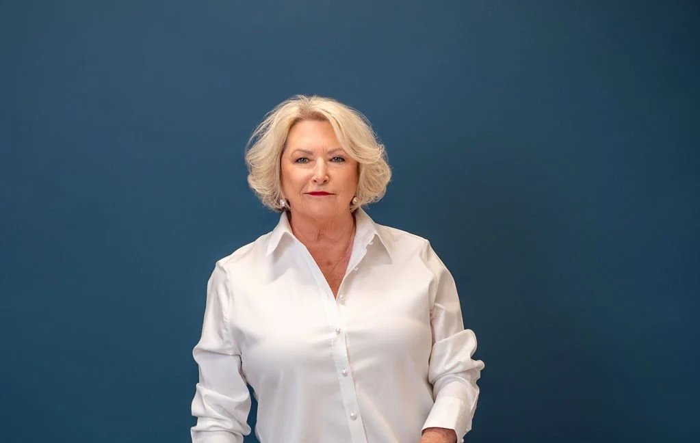 An older woman with blonde, wavy hair wearing a white button-up shirt against a plain blue background.