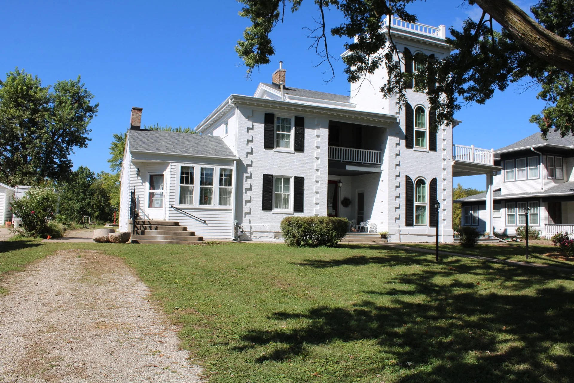 The Downing House - A white, multi-story house with black window shutters, a small side porch, and a larger front porch, surrounded by a well-maintained lawn and a gravel driveway, with trees and neighboring houses in the background.