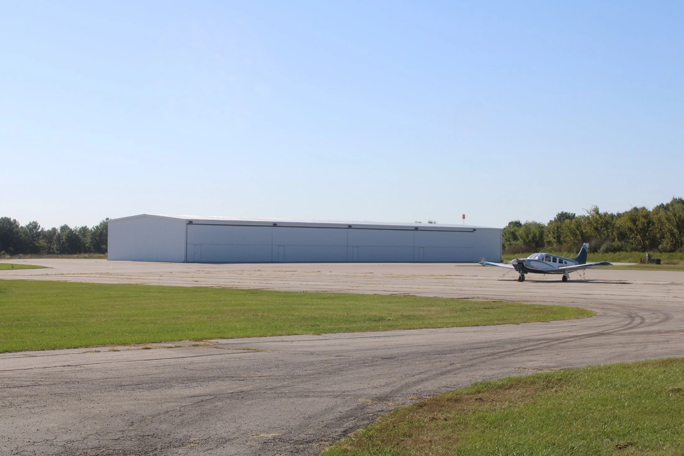 Small aircraft parked on a tarmac with a large white hangar building in the background, green grass in the foreground, and trees and a clear blue sky behind.