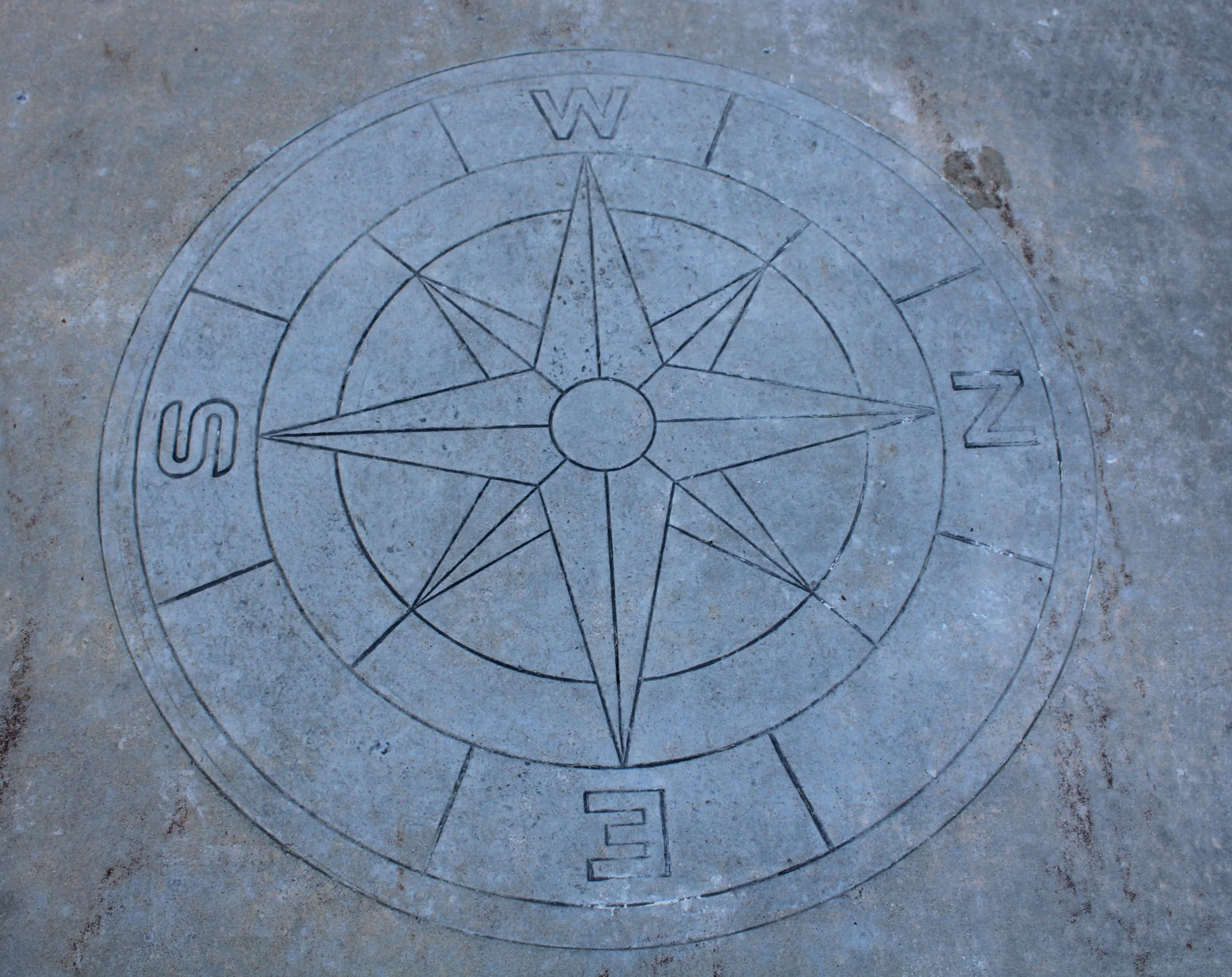 A compass rose engraved on a concrete surface showing the cardinal directions North, South, East, and West.