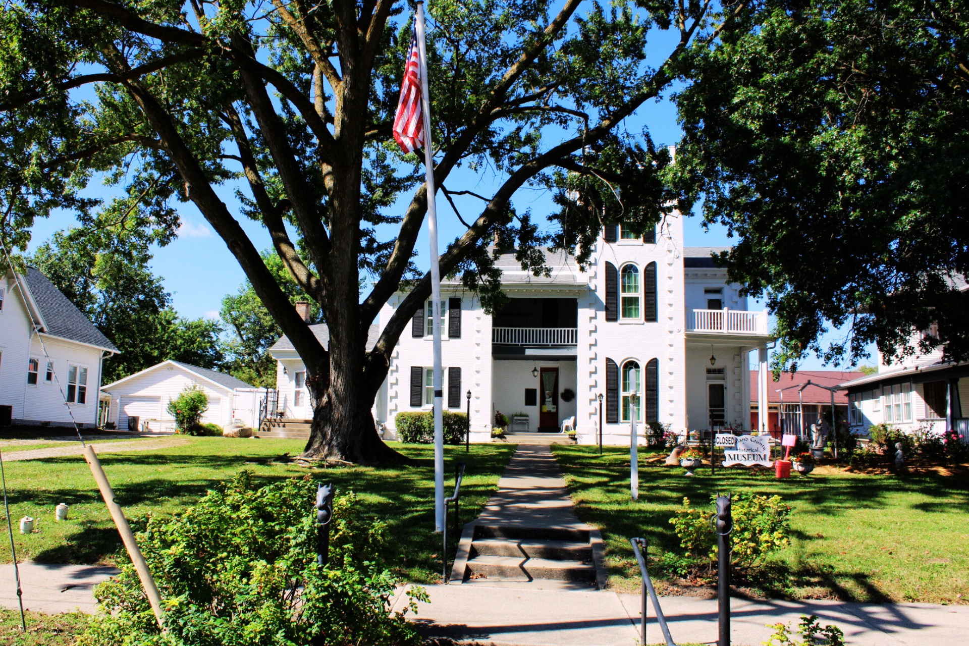 The Downing House - White building with black shutters, large trees, green lawn, American flag, and a sign indicating a historical museum.