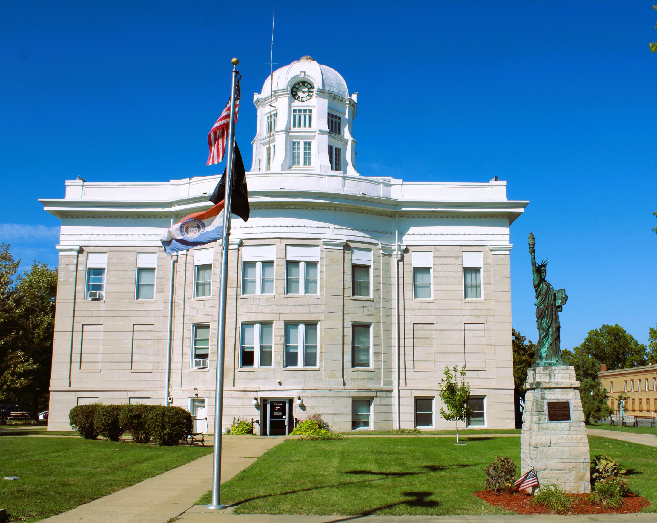 A historic government building with a clock tower, flags, a statue of the Statue of Liberty, and a well-maintained lawn under a clear blue sky.