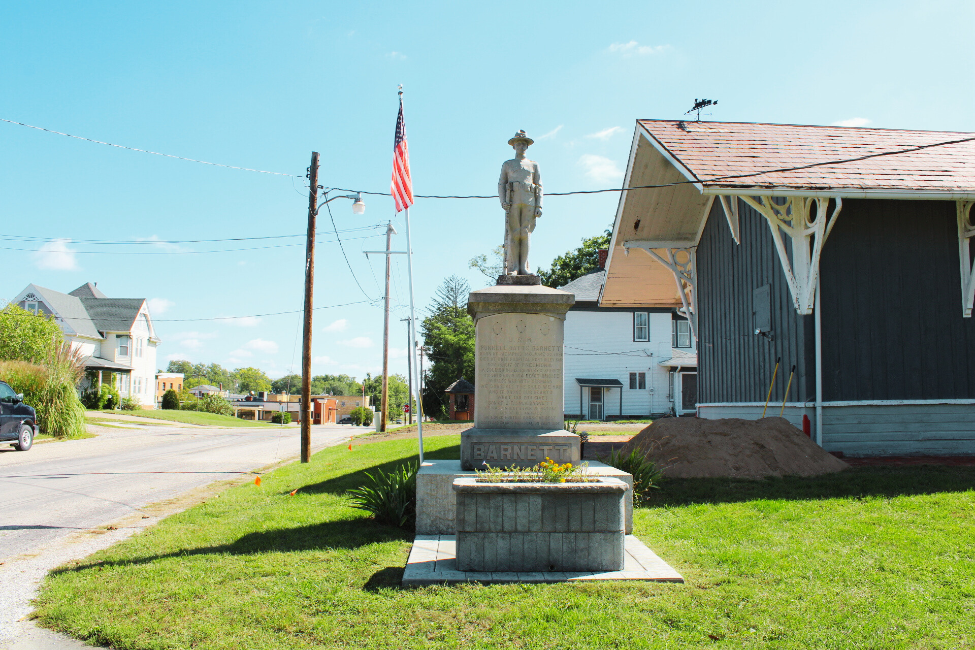 A war memorial monument with a statue of a soldier on top, located on a grassy area next to the road, with an American flag and a weather vane on nearby buildings.
