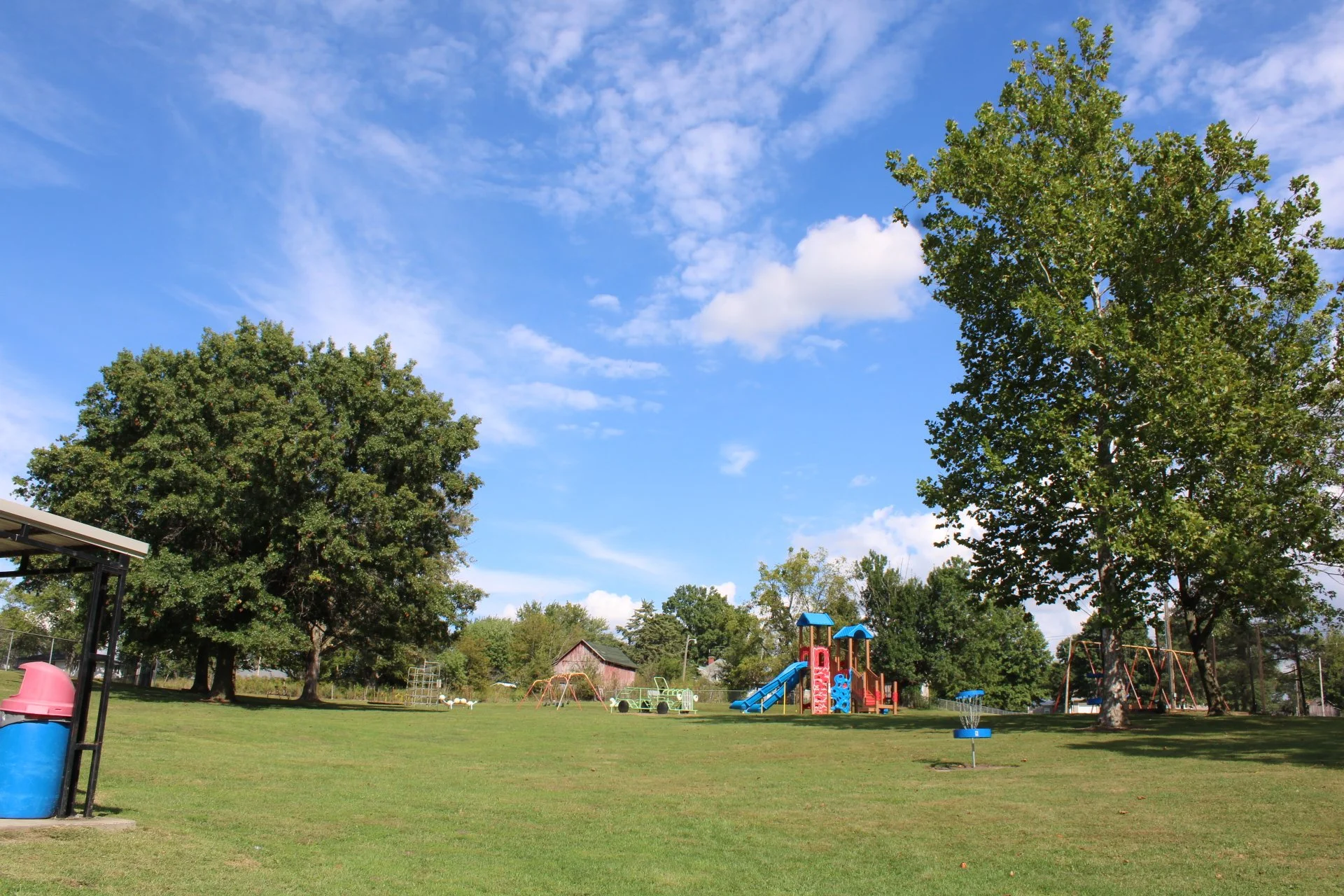 A park with green grass, tall trees, playground equipment like slides and swings, and a blue sky with scattered clouds.
