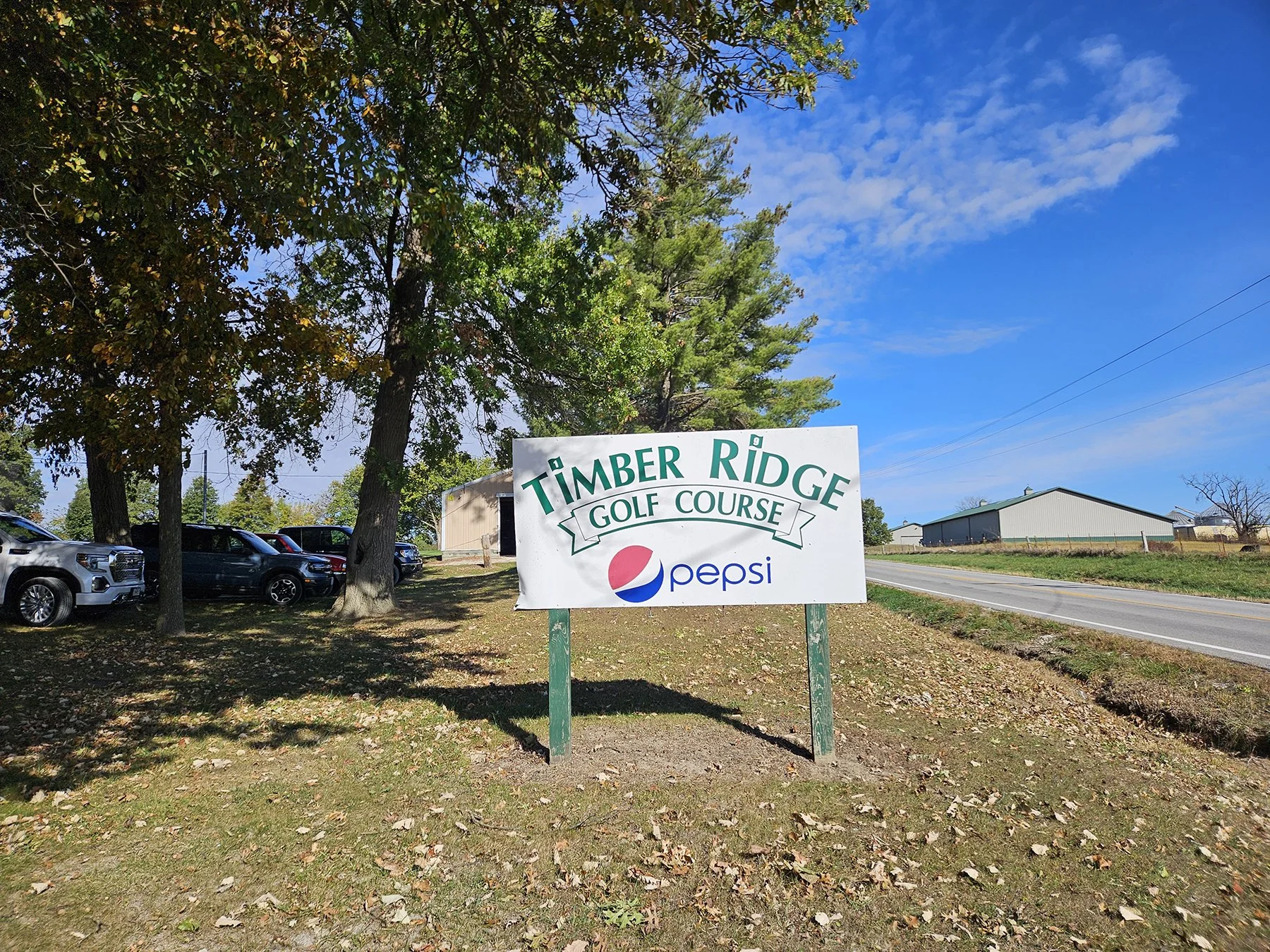 A sign for Timber Ridge Golf Course with the Pepsi logo beneath, situated outdoors under trees on a partly cloudy day, with a parking lot of cars and a road nearby.