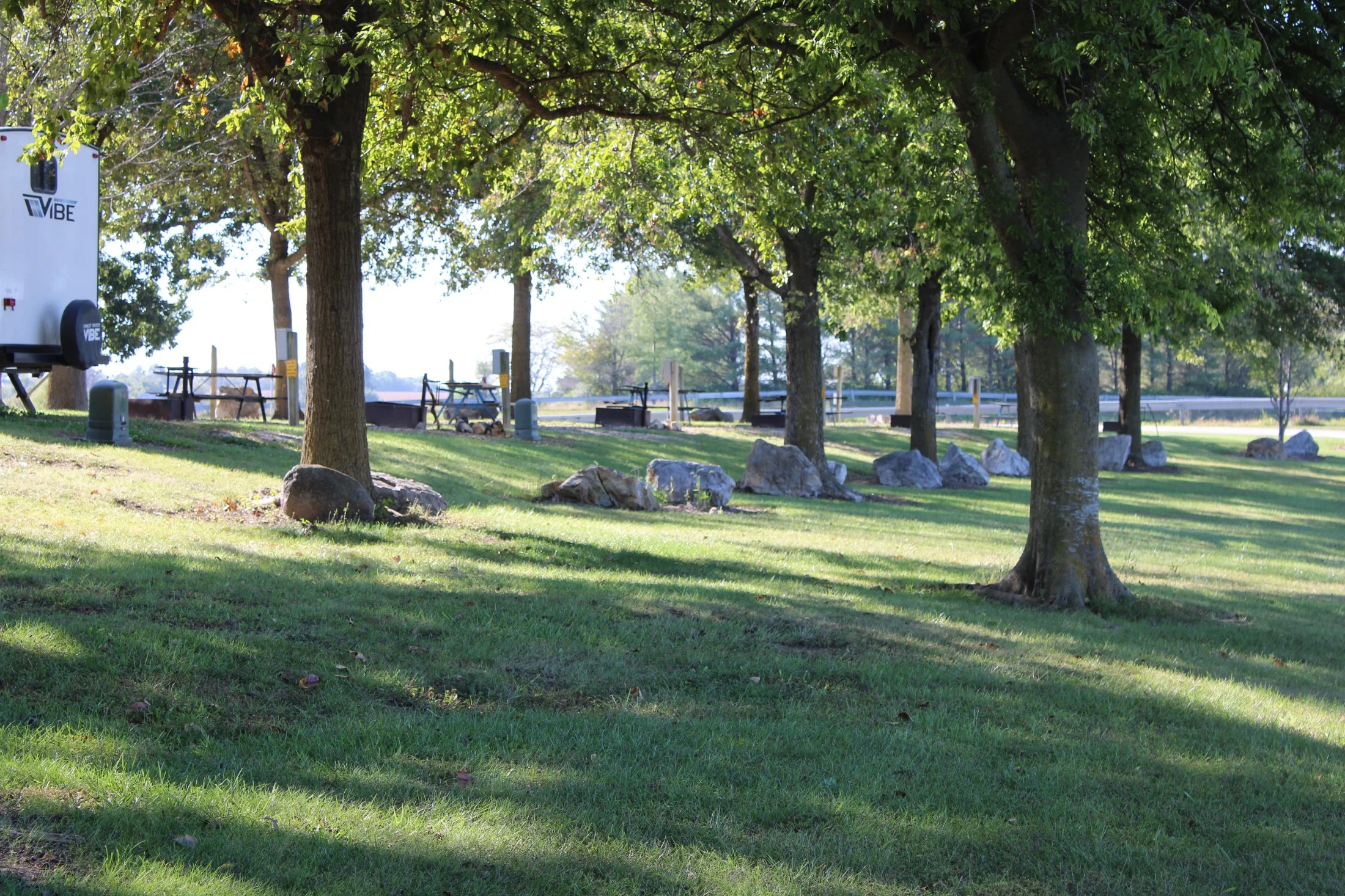 A park with green grass, tall trees, and large rocks, with picnic tables, a trailer, and a fence in the background.
