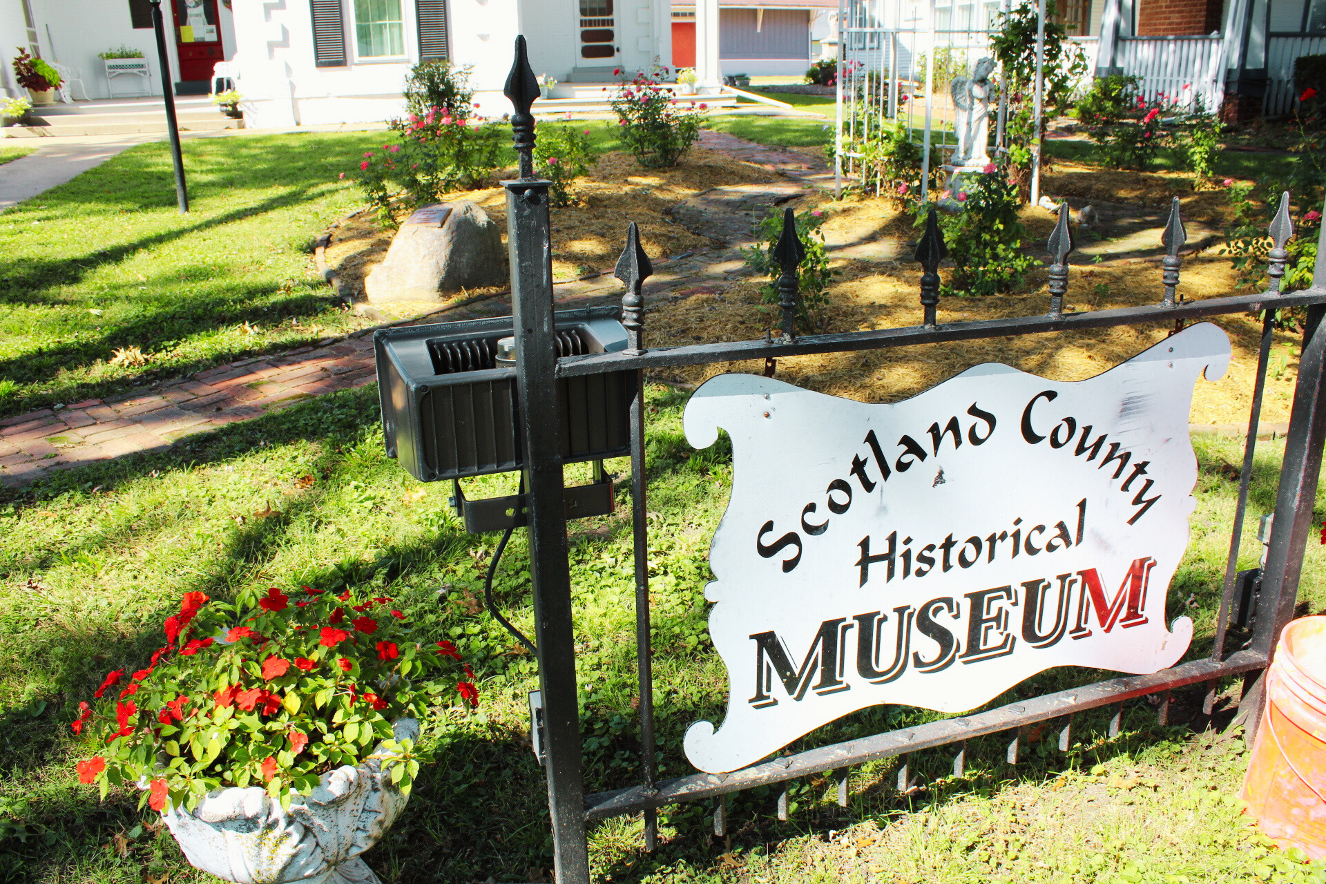 Entrance sign for Scotlans County Historical Museum, with a flower planter nearby, in a garden with roses and a brick walkway