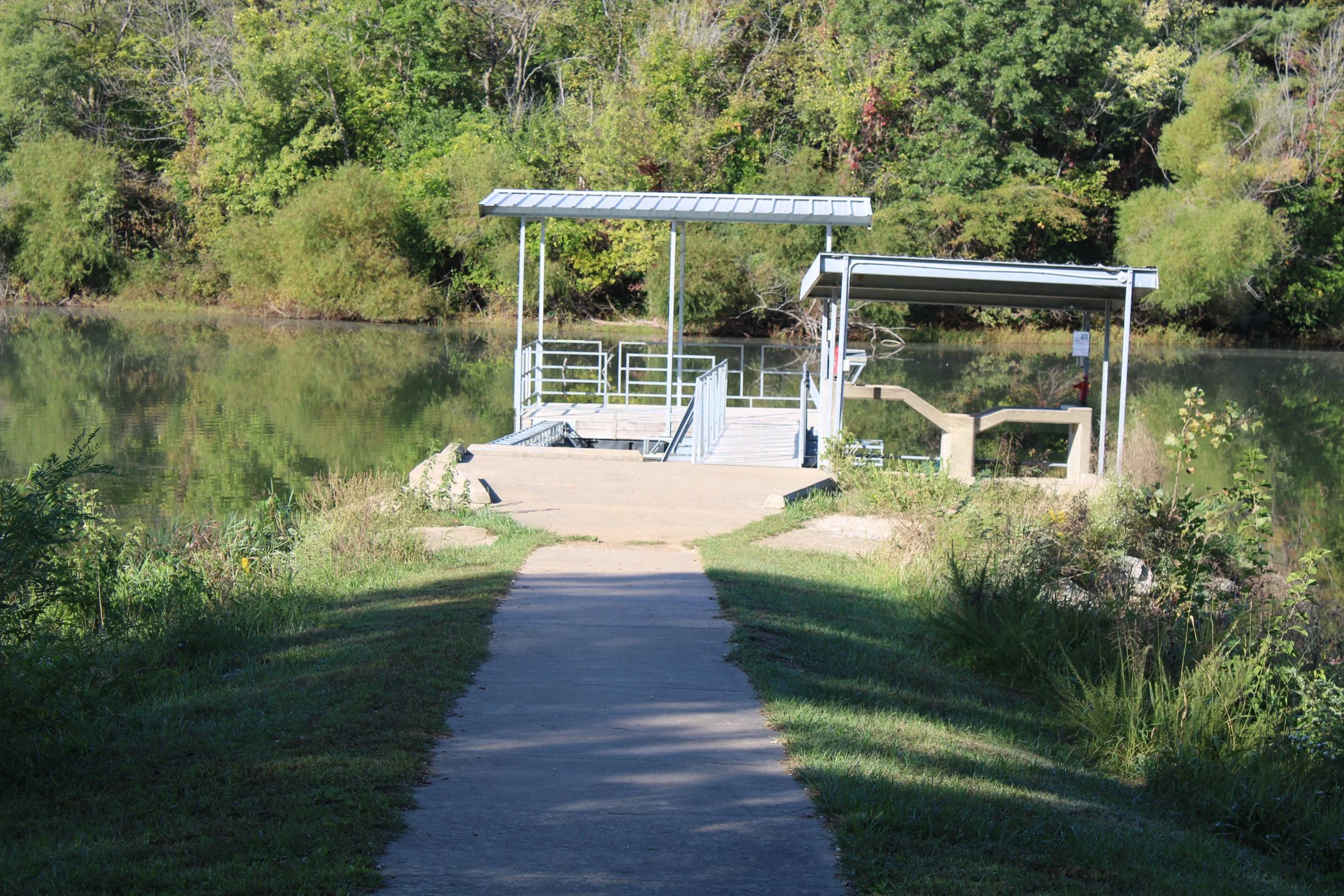 Path leading to a boat launch area with covered structures, railing, and stairs, situated near calm water with green trees in the background.