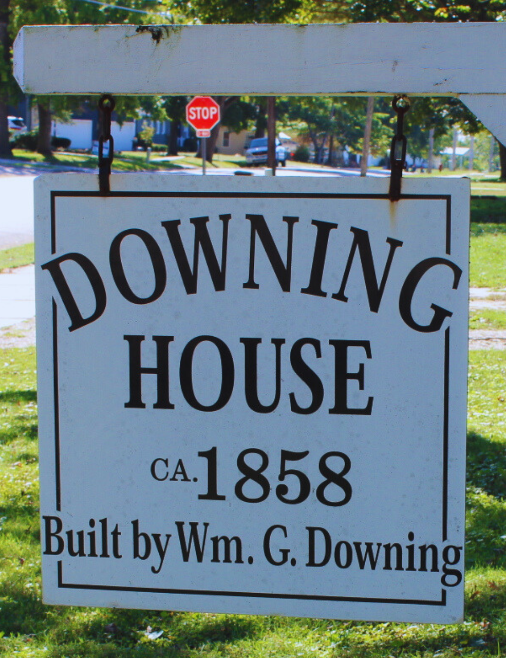 Historical house sign that reads 'Downing House, ca. 1858, built by Wm. G. Downing, hanging on a white wooden frame with trees and houses in the background.'