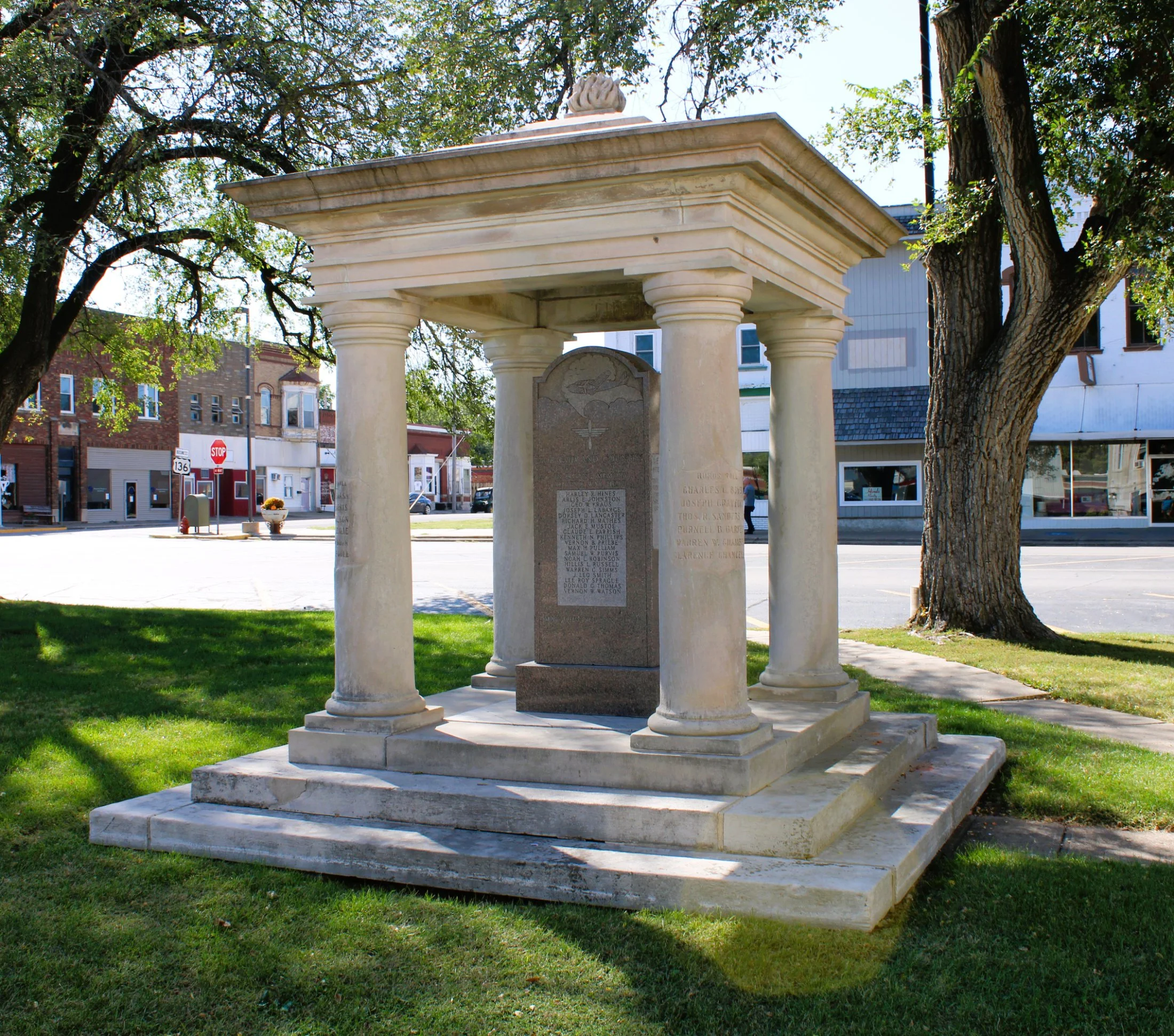A monument with four columns and a capped roof, situated on a grassy area next to a sidewalk and street, with buildings and trees in the background.