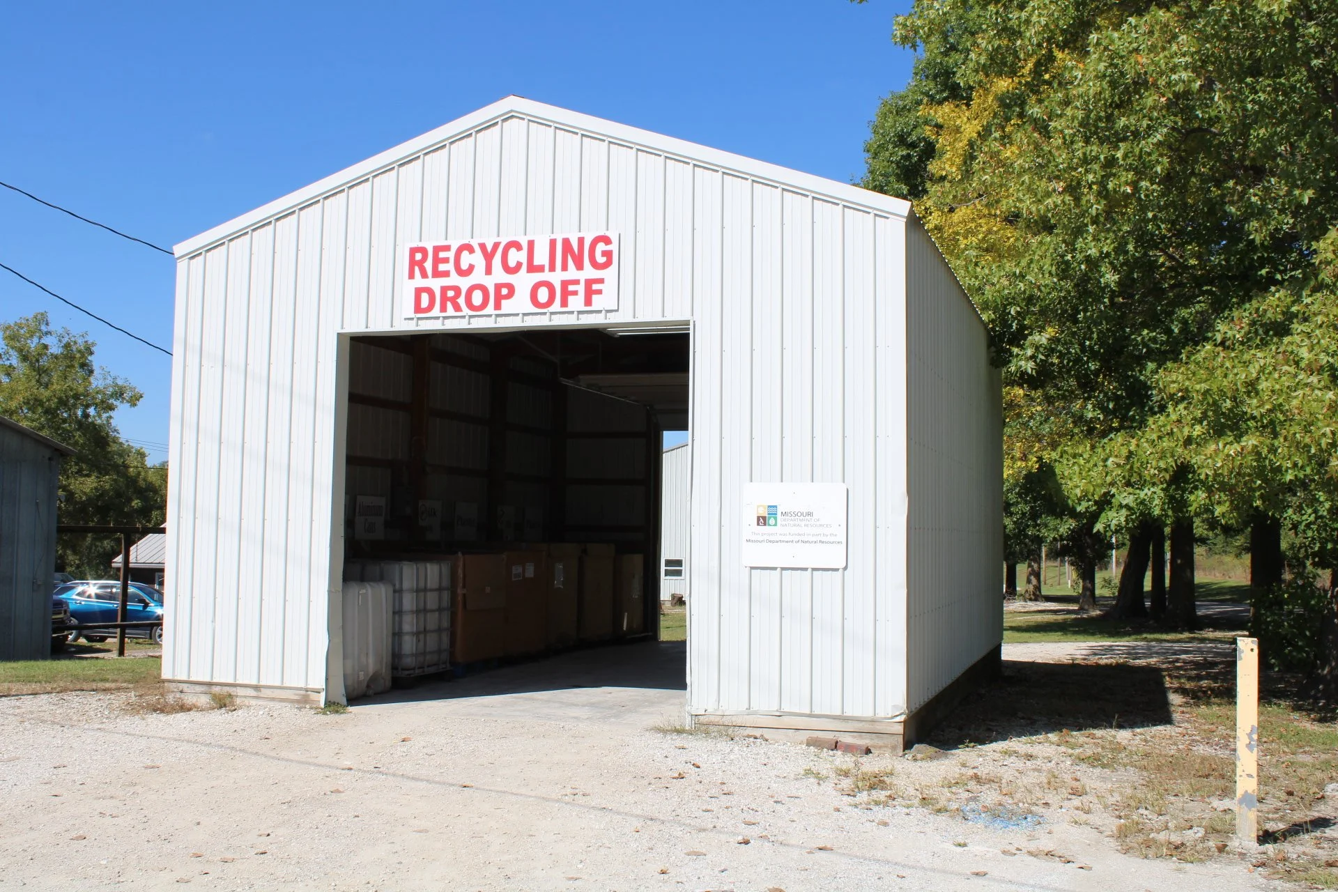 White metal shed with a sign that reads 'Recycling Drop Off' in red letters, surrounded by trees and a parking lot in the background.