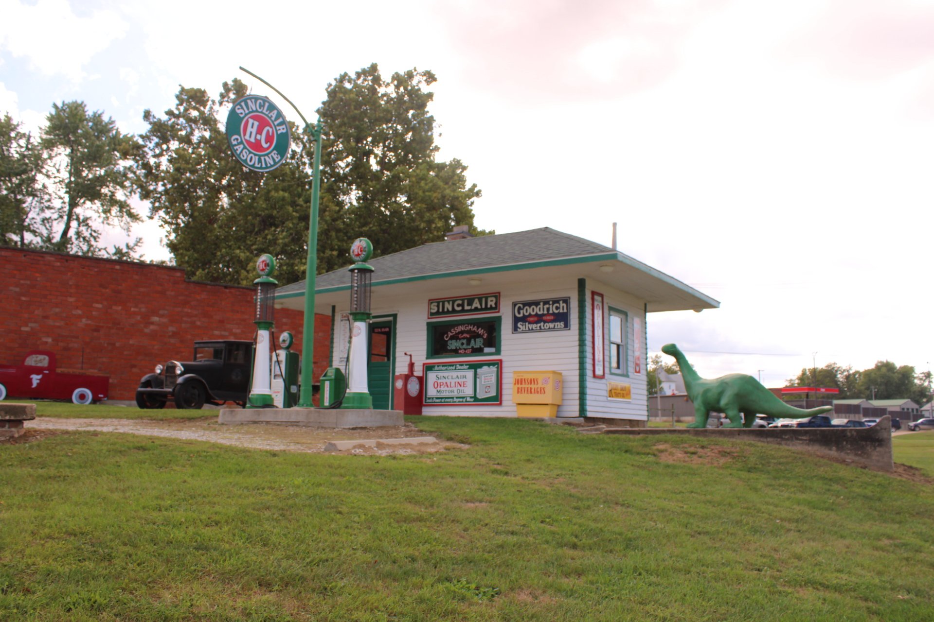 Vintage gas station with Sinclair brand signs, old fuel pumps, a toy dinosaur statue, and vintage cars parked nearby.