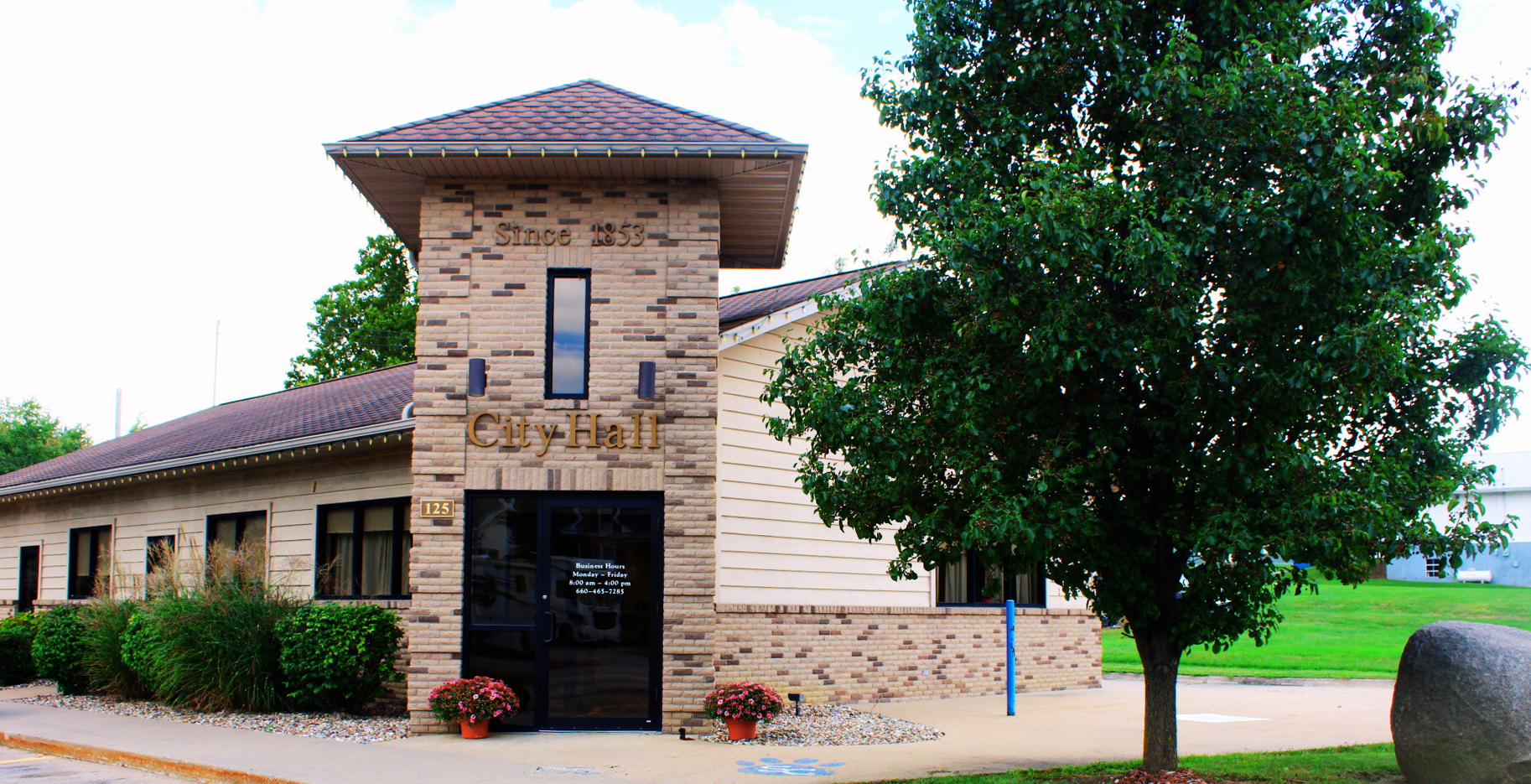 Town hall building with brick tower, sign that reads 'City Hall,' and trees in front, during daytime.