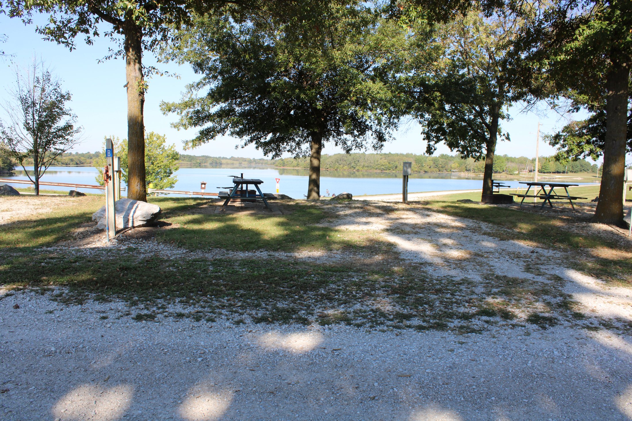 A peaceful lakeside scene with trees, picnic tables, and a gravel pathway.
