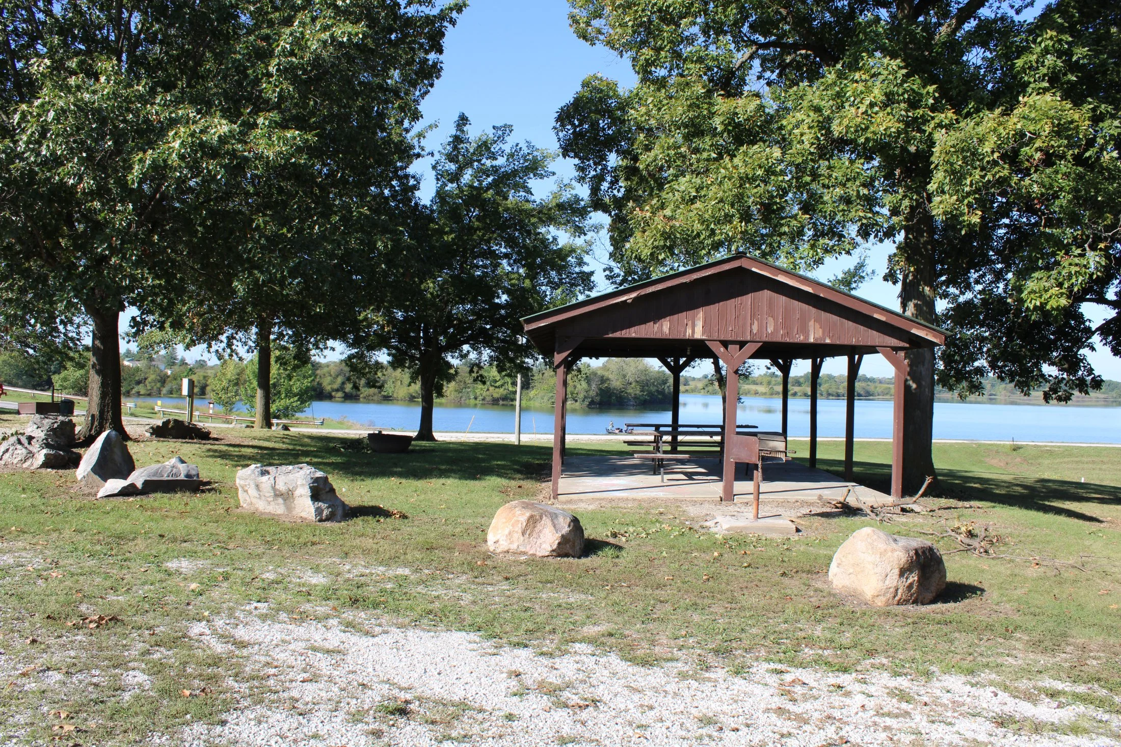 A park with a wooden picnic shelter, trees, rocks, and a view of a lake in the background under a clear blue sky.