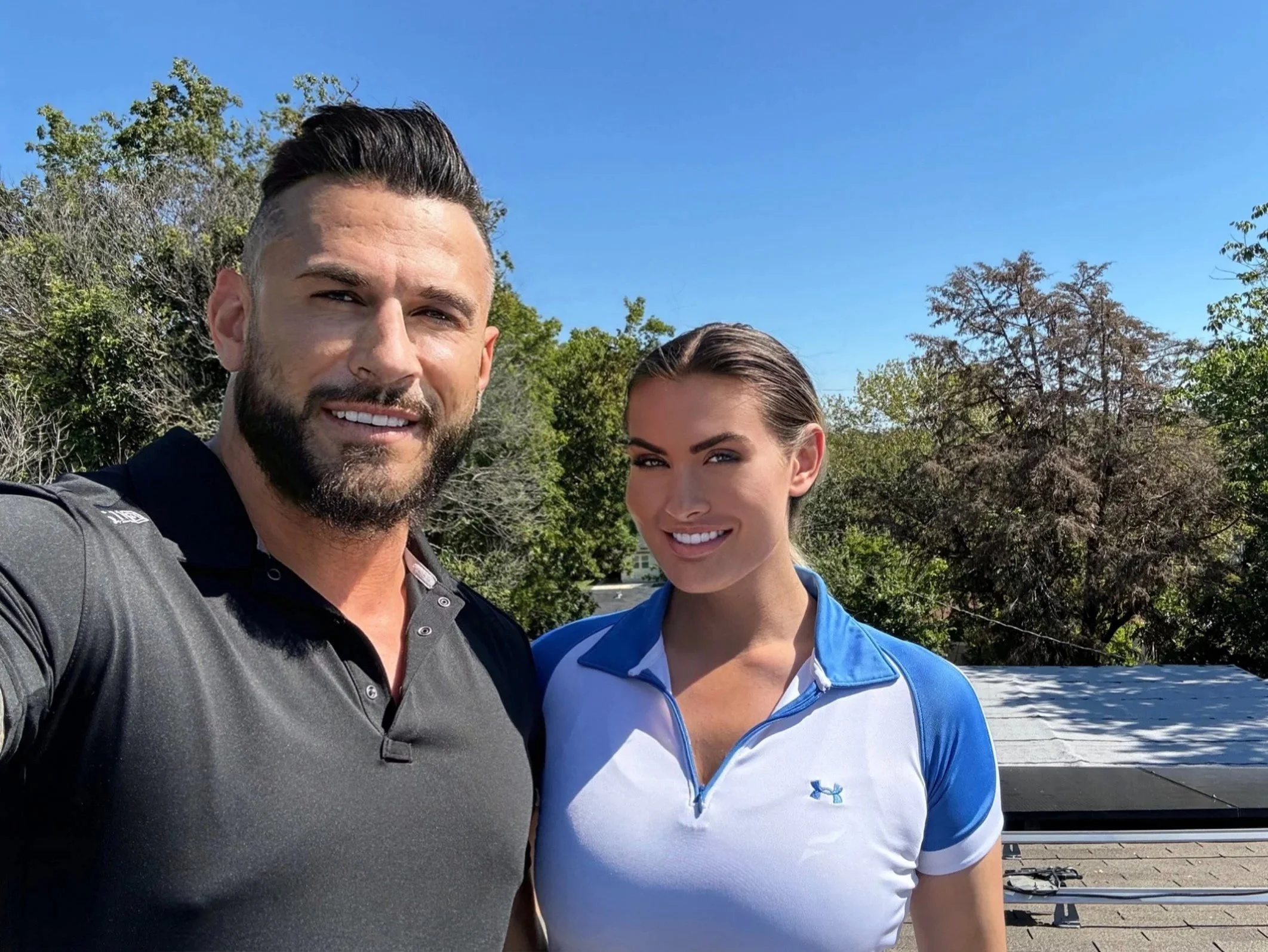 A man and a woman taking a selfie outdoors on a sunny day with blue sky and green trees in the background.