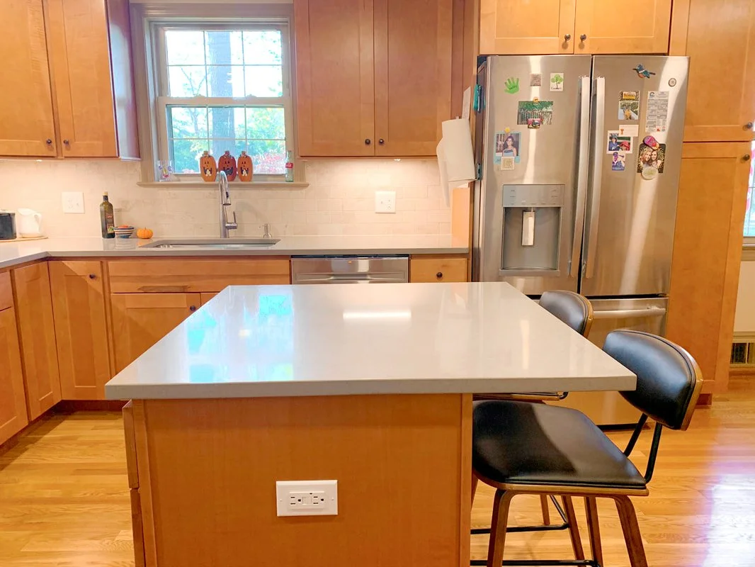 Kitchen with wooden cabinets, a white countertop island, stainless steel refrigerator with photos and stickers, a double sink under a window, and two black barstools by the island.