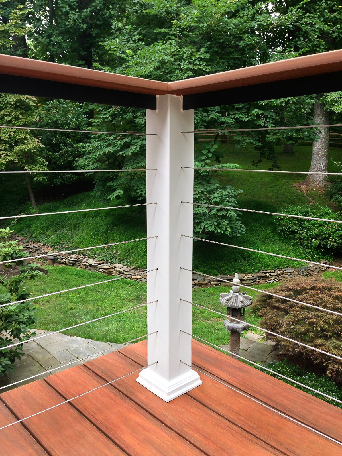 View from a wooden deck with cable railing, overlooking a lush green backyard with trees, a stone pathway, a Japanese-style lantern, and dense foliage.
