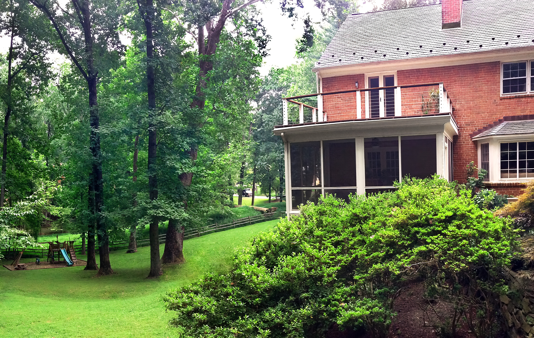 A backyard with green grass, tall trees, a small playground setup, and a red brick house with a screened porch and a balcony.